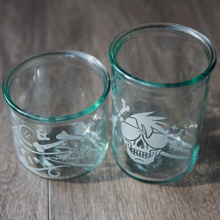 Two straight-sided drinking glasses with a slight green-blue tinge, viewed slightly from above. One is shorter and has a hand and assorted bones on it. The second glass is taller and has a skull with a mohawk wearing sunglasses with a sword and bones behind it.