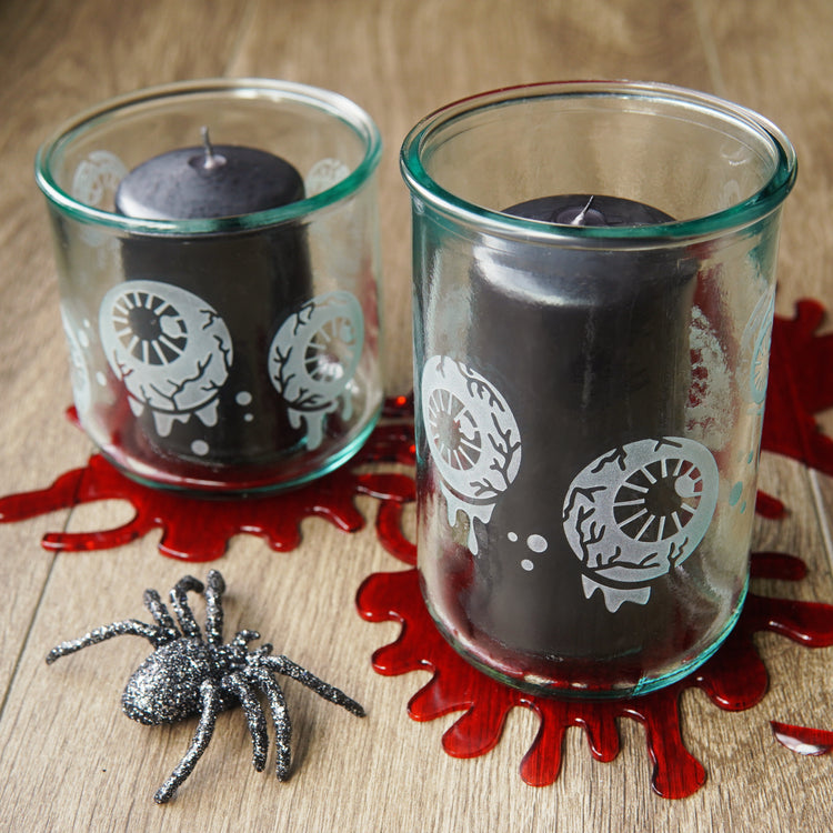 dripping eyeballs etched onto 2 different sized green-tinged glass cups holding black pillar candles. They're sitting on bright red "blood" splatters, next to a glitter-encrusted spider