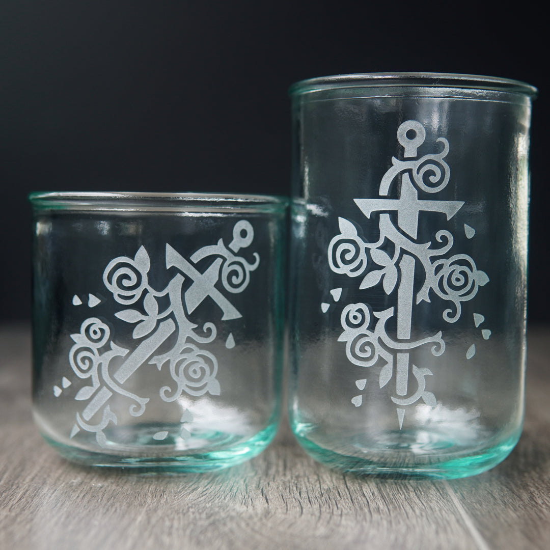 Sword wrapped with a rose vine engraved onto short and tall drinking glasses. The glass has a slight bluegreen tinge.