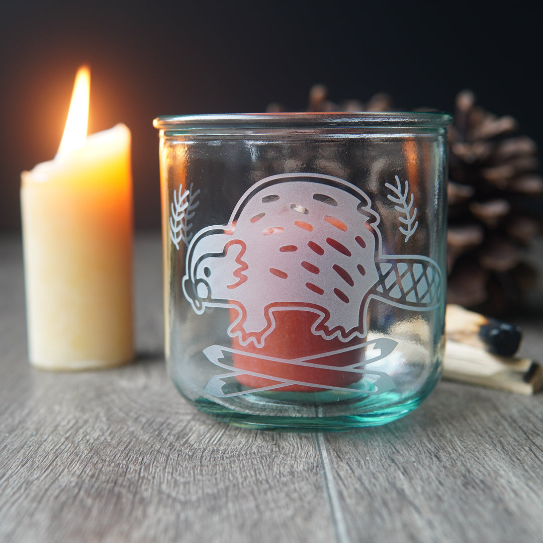 short recycled glass engraved with a cute beaver, with a red votive candle inside it. A lit candle is beside it.