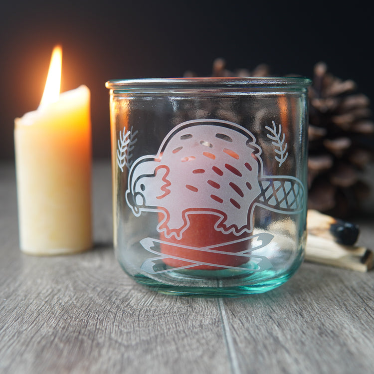 short recycled glass engraved with a cute beaver, with a red votive candle inside it. A lit candle is beside it.