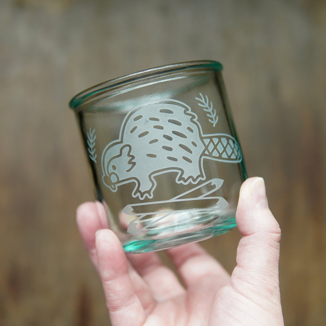 short recycled glass cup engraved with a cute beaver and crossed logs, held in a hand. The glass has a green-blue tinge.
