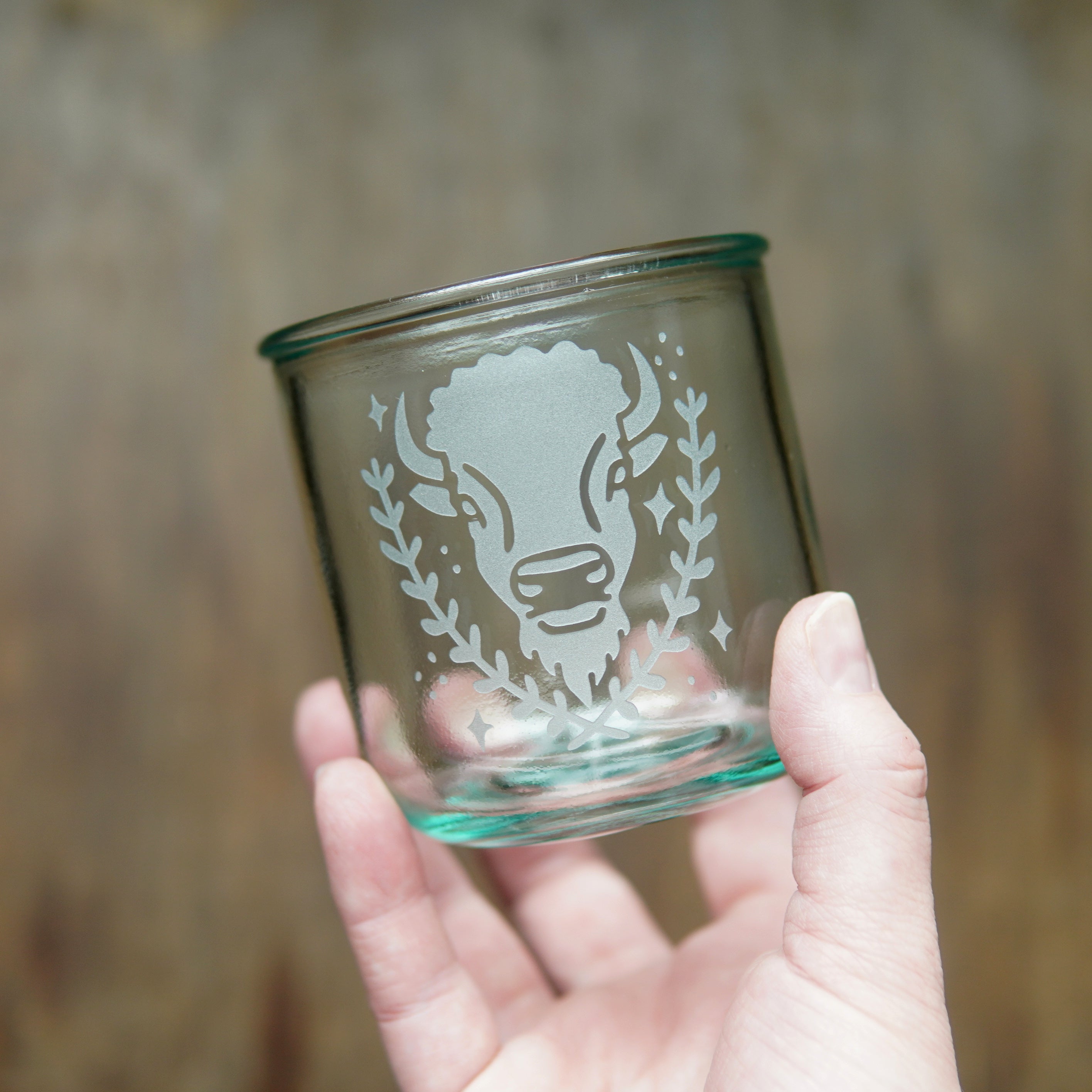 Bison engraved on a short blue-tinged recycled glass cup, held in a hand