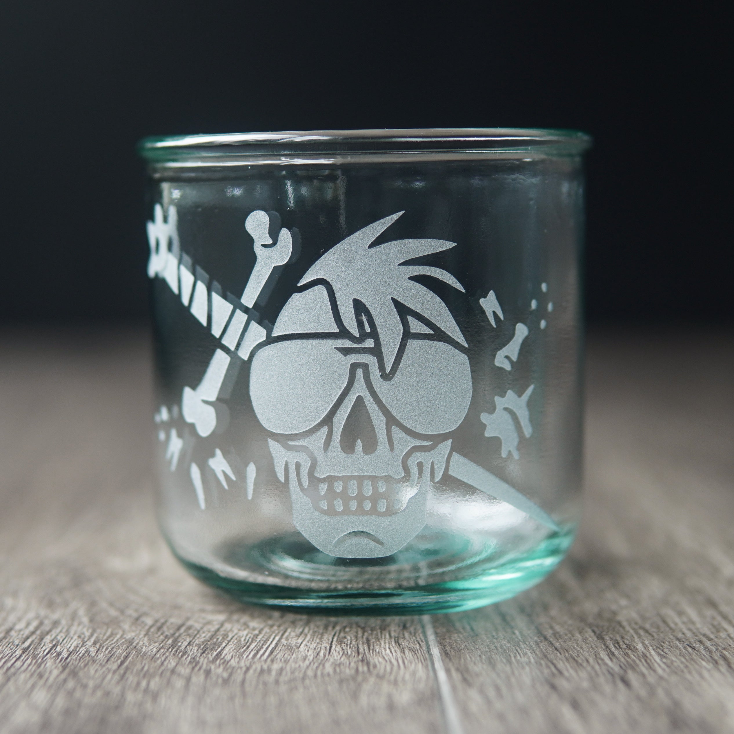 A straight-sided lowball glass with a slight green-blue tinge. The glass has a skull with a mohawk wearing sunglasses, with a sword and bones behind it.