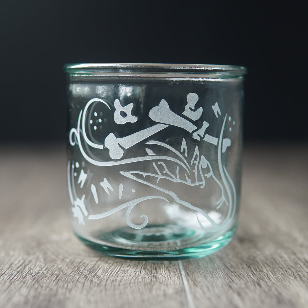 A straight-sided lowball glass with a slight green-blue tinge. The glass has  a hand and assorted bones on it, with swirly spirals suggesting magic and movement between the hand and bones.