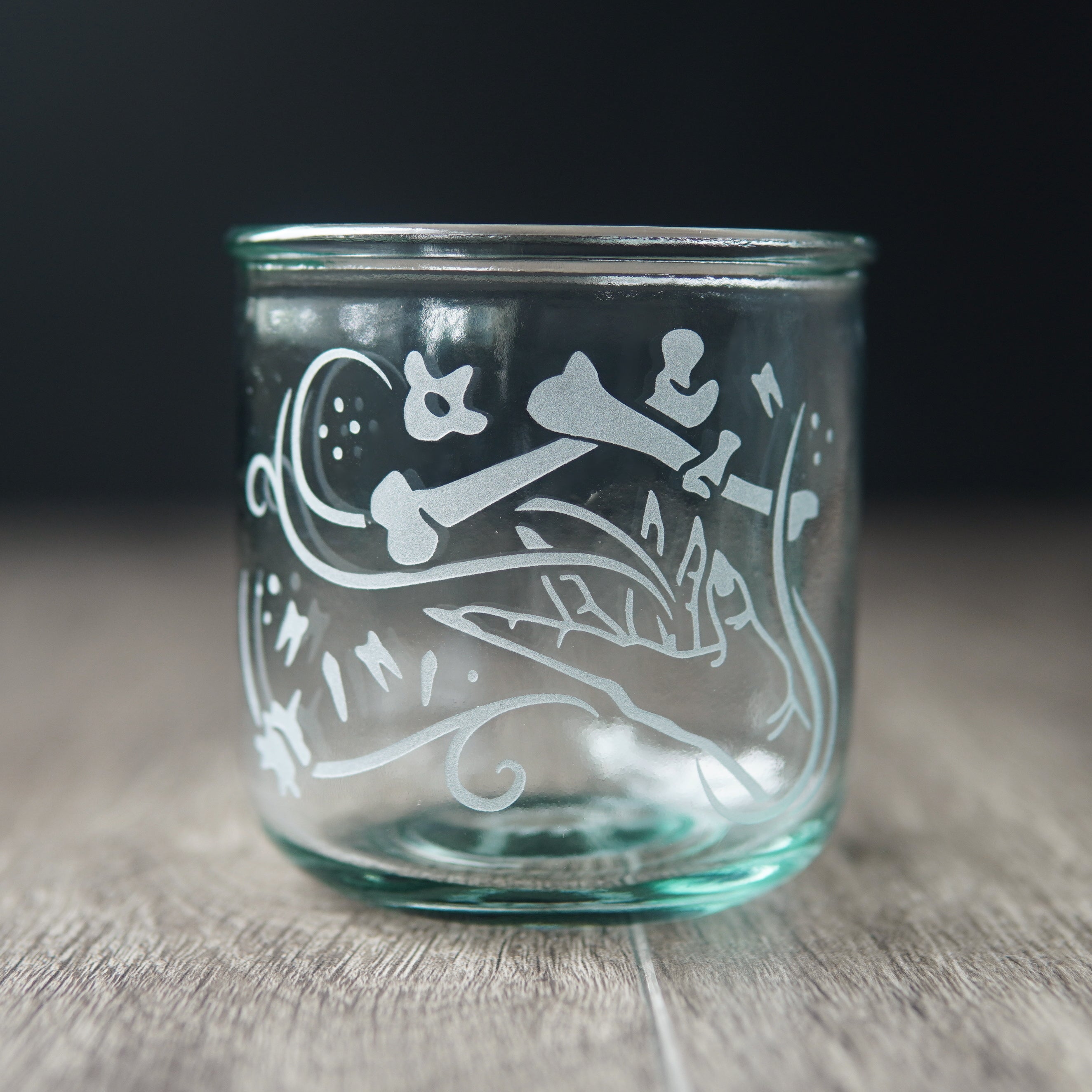 A straight-sided lowball glass with a slight green-blue tinge. The glass has  a hand and assorted bones on it, with swirly spirals suggesting magic and movement between the hand and bones.