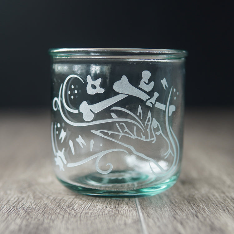 A straight-sided lowball glass with a slight green-blue tinge. The glass has  a hand and assorted bones on it, with swirly spirals suggesting magic and movement between the hand and bones.