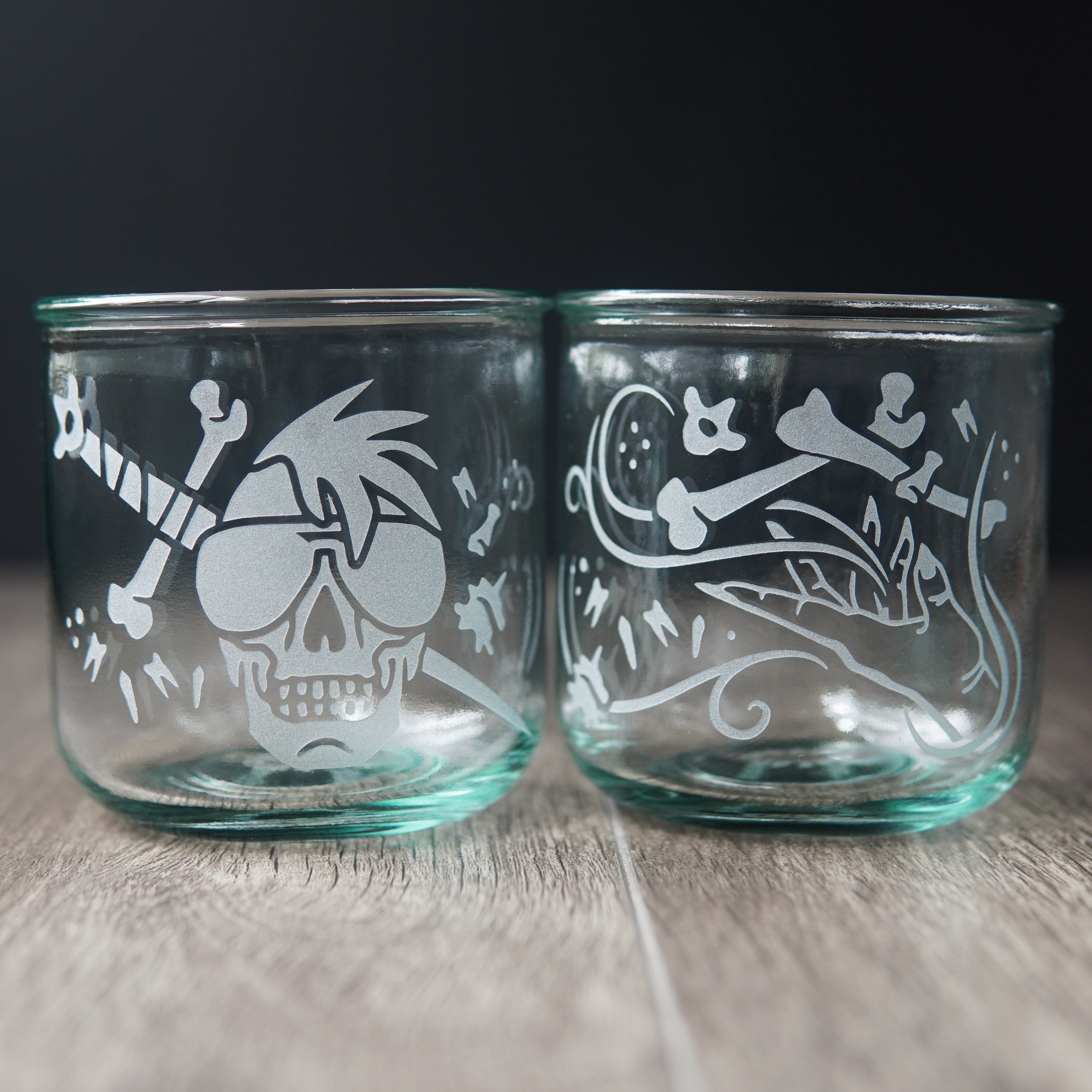 Two straight-sided drinking glasses with a slight green-blue tinge. One has a hand and assorted bones on it. The second glass has a skull with a mohawk wearing sunglasses with a sword and bones behind it.