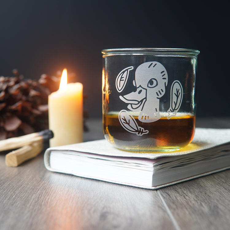 Duck engraved short glass of whiskey, sitting on a book next to a lit candle, palo santo, and pine cones