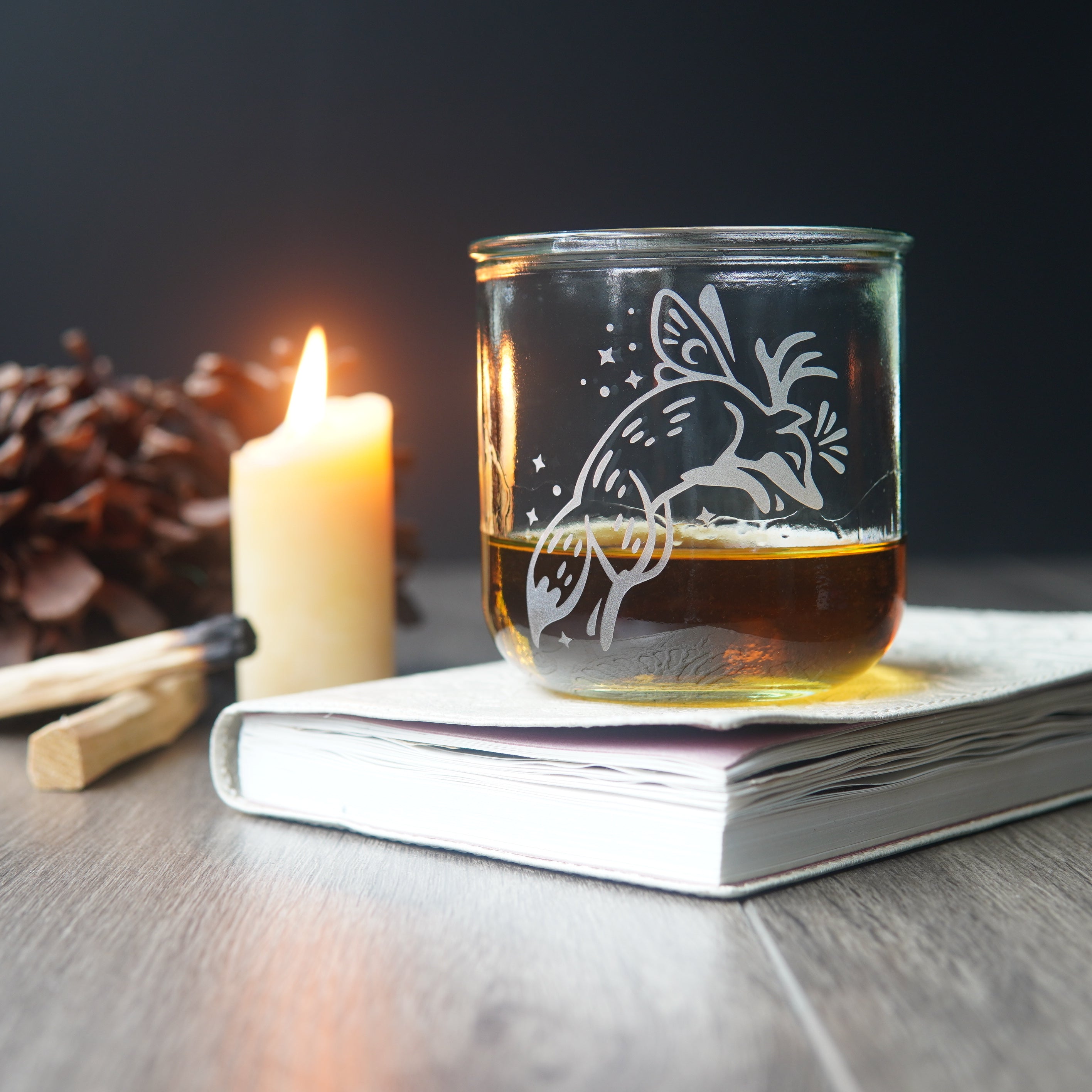 Fox with fairy wings etched onto a short glass, sitting on a book next to a lit candle, pine cones, and palo santo
