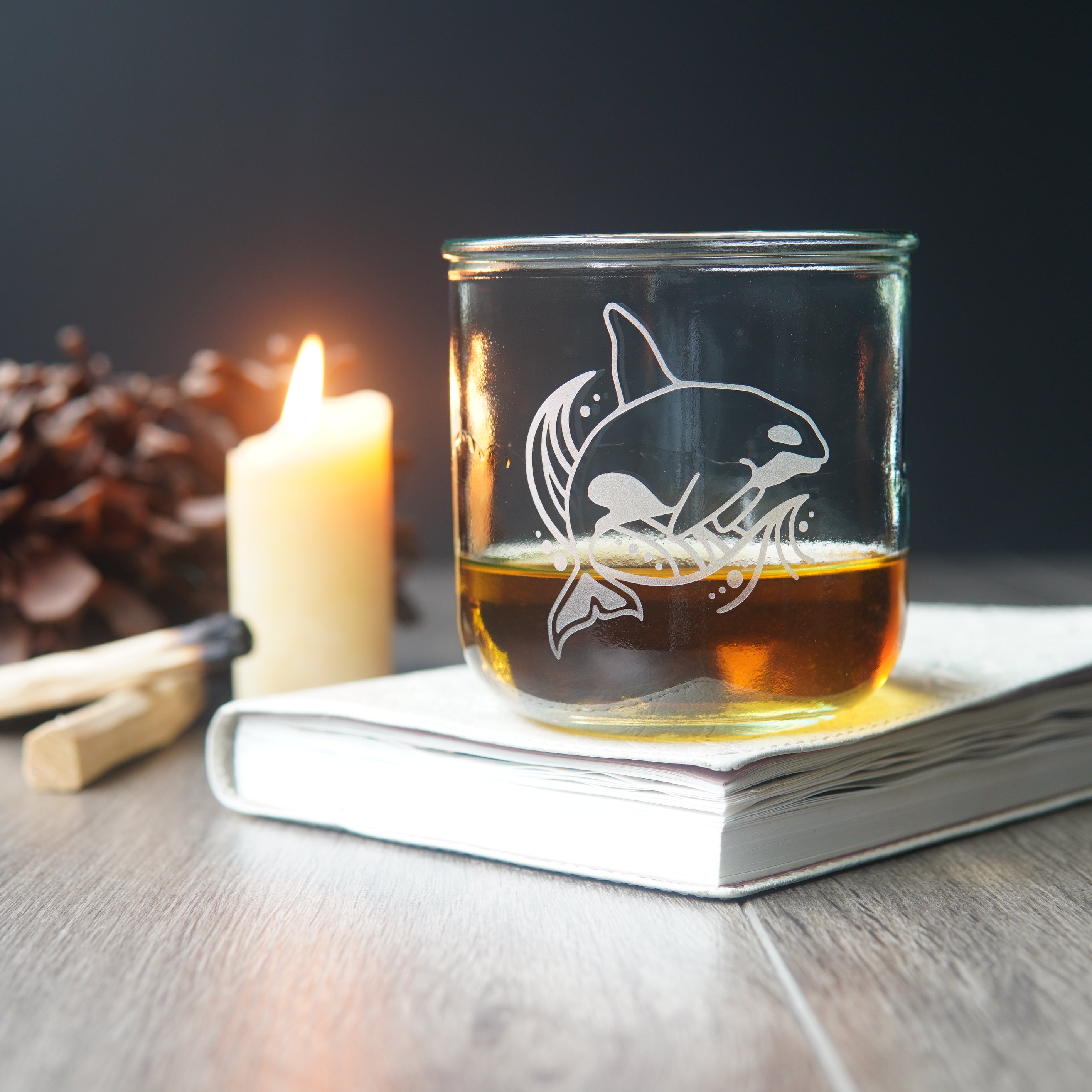 killer whale etched onto a short glass of whiskey beside a book and lit candle