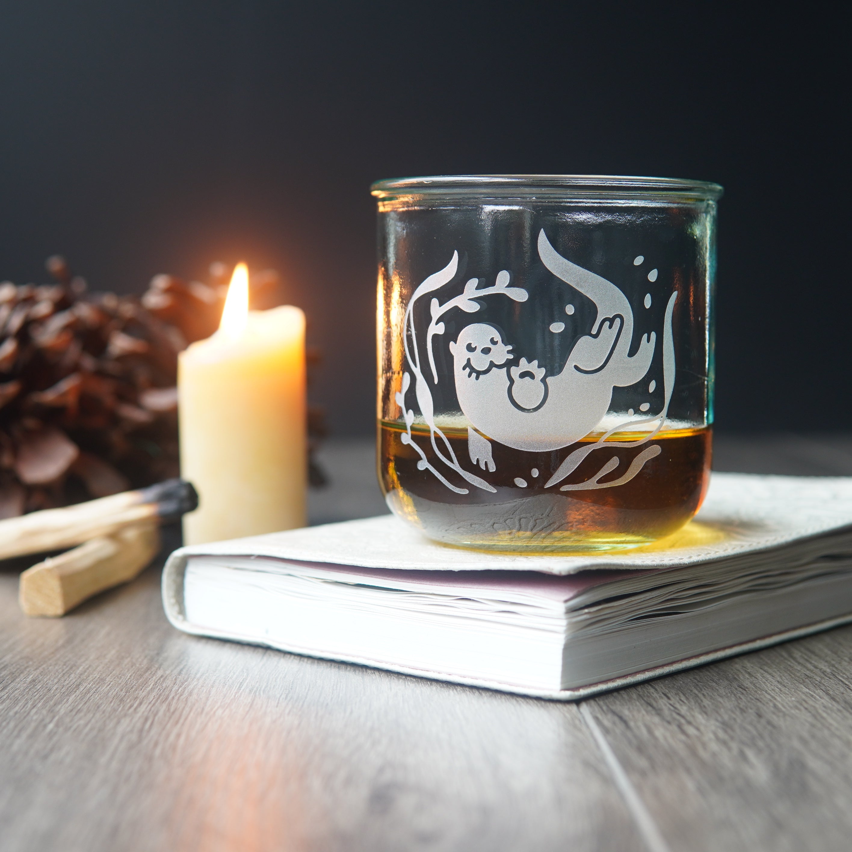 short otter glass of whiskey, next to a lit candle and book