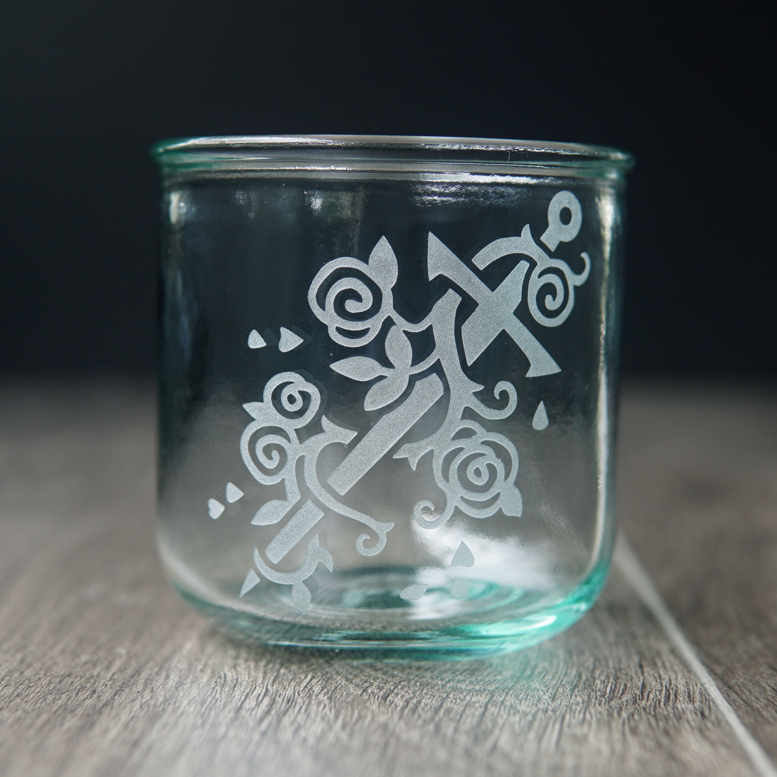 Sword wrapped with a rose vine engraved onto a short drinking glass. The glass has a slight bluegreen tinge.