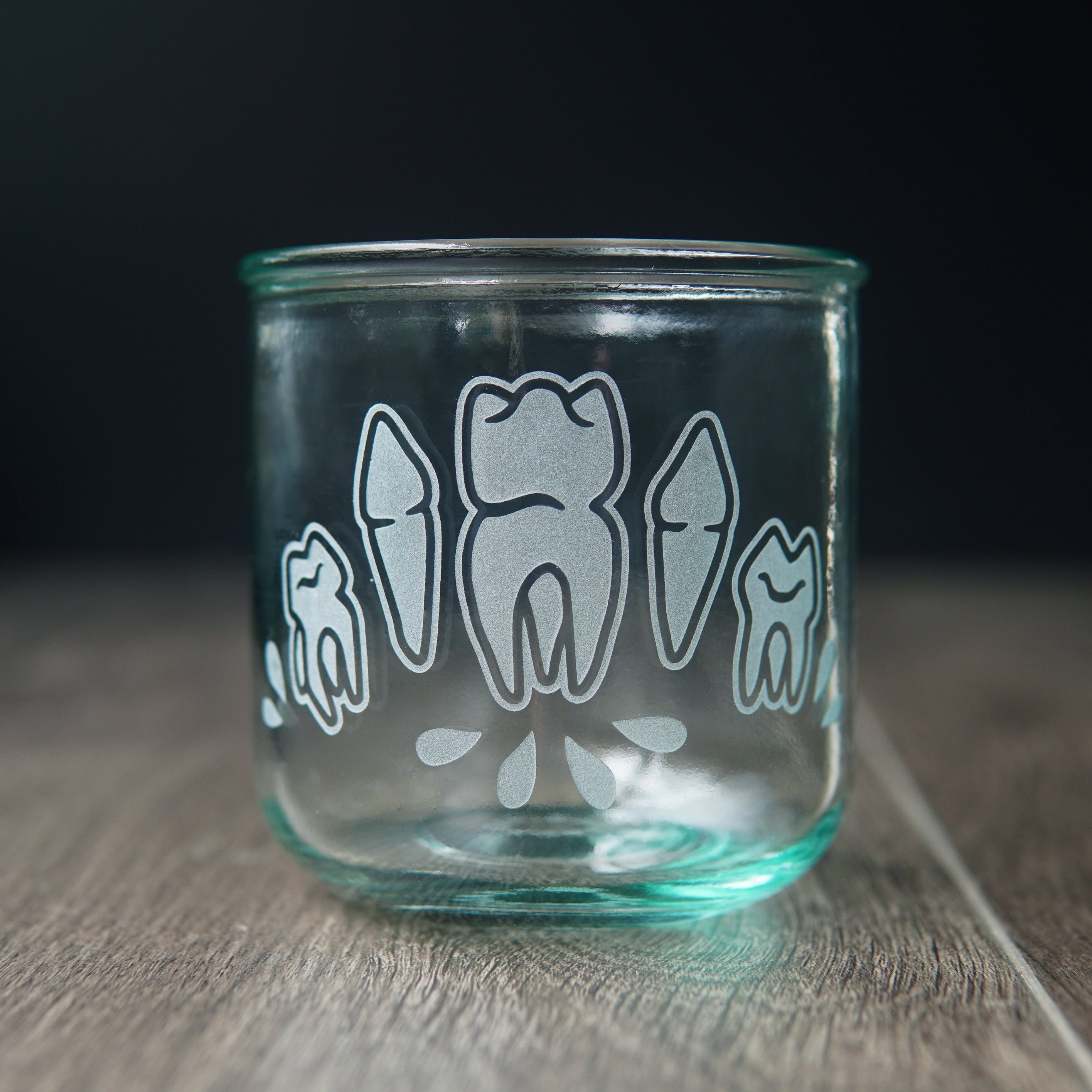 Four teeth with blood droplets engraved onto a short drinking glass. The glass has a slight bluegreen tinge.