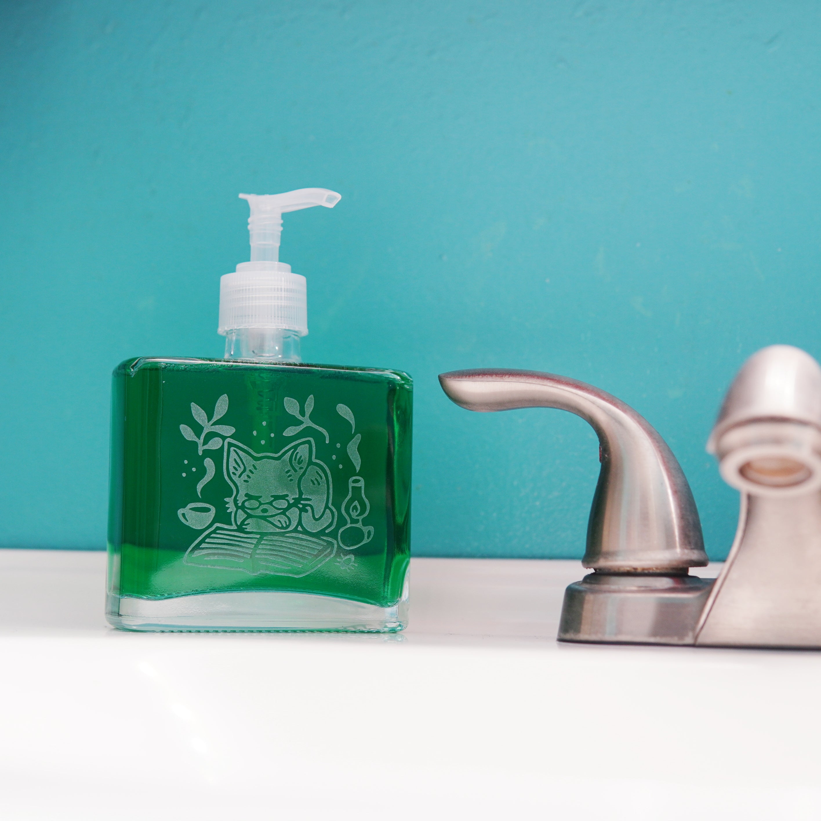 glass pump bottle soap dispenser full of green soap next to a bathroom faucet, with a cat sleeping on an open book engraved on it