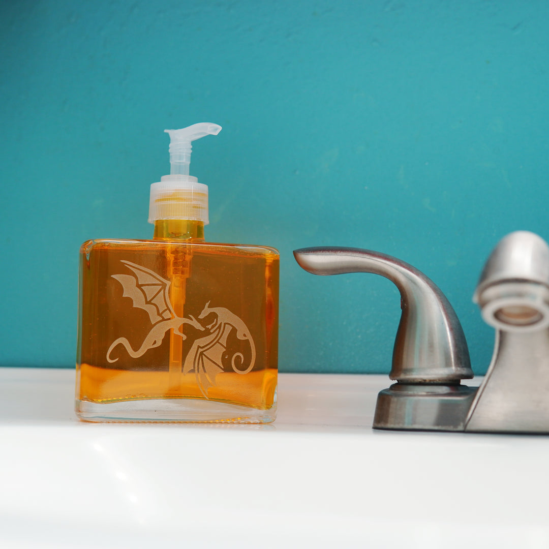 glass pump bottle soap dispenser full of orange soap next to a bathroom faucet, with a pair of dragons engraved on it