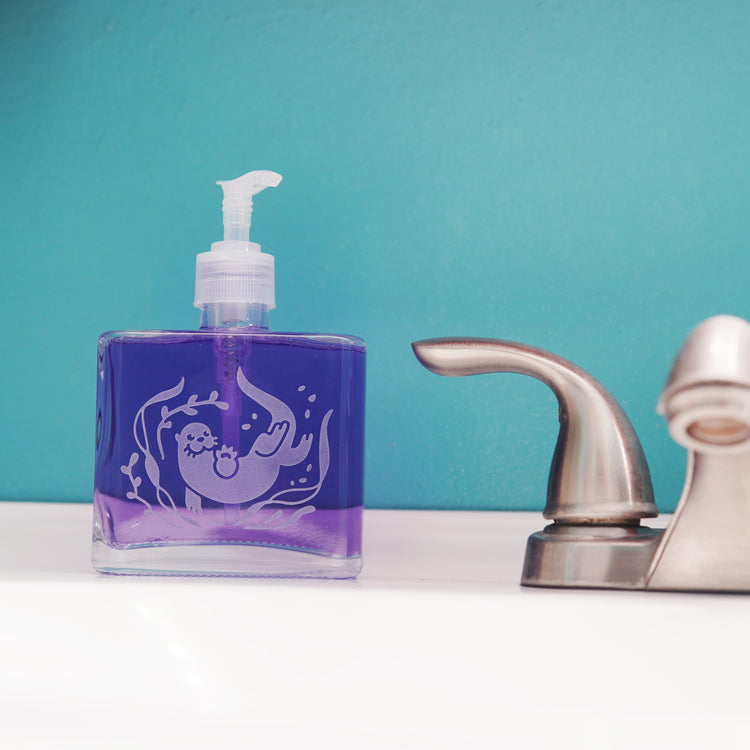 glass pump bottle soap dispenser full of purple soap next to a bathroom faucet, with an otter engraved on it