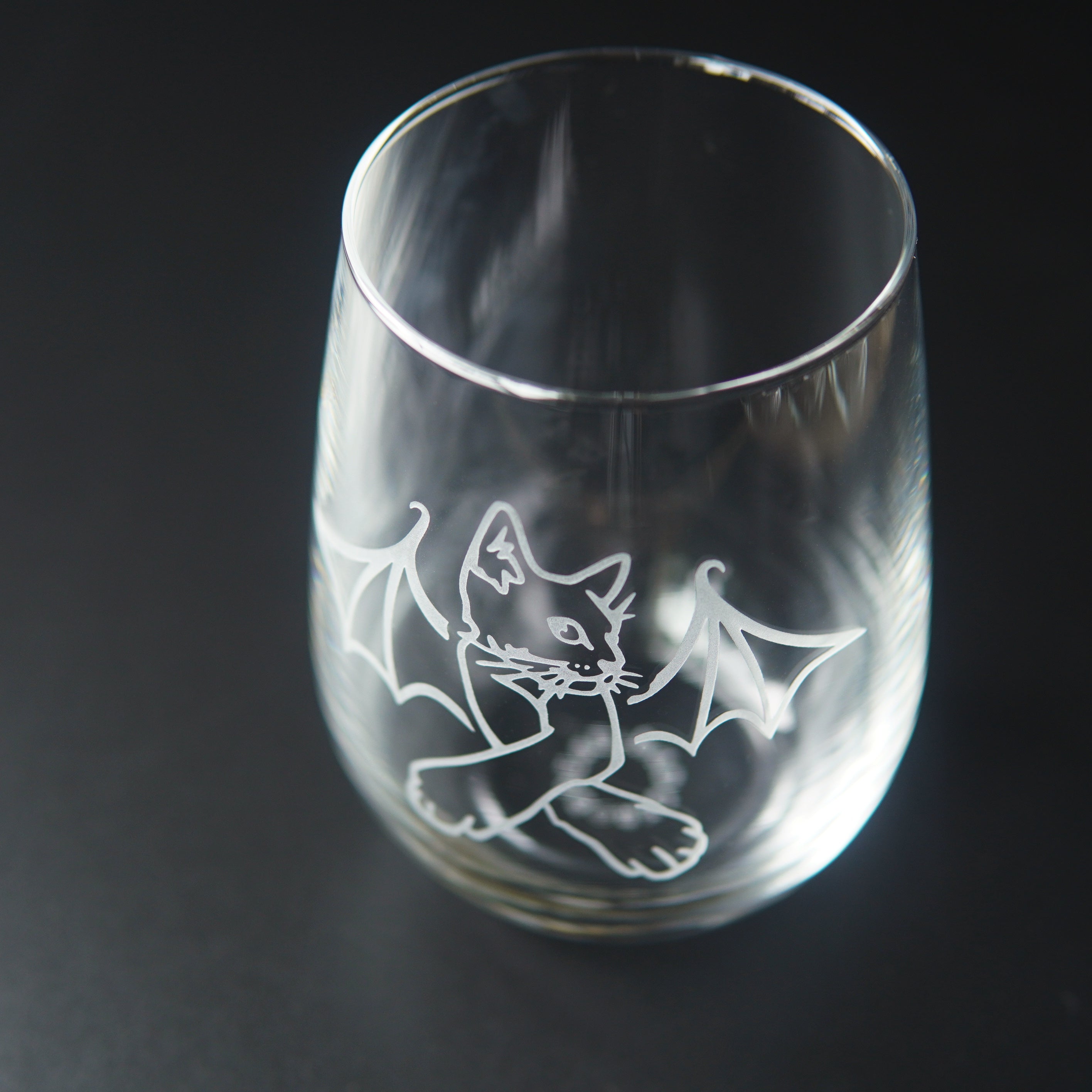 stemless wine glass engraved with a cat with crossed arms and bat wings. We're viewing the glass mostly from above