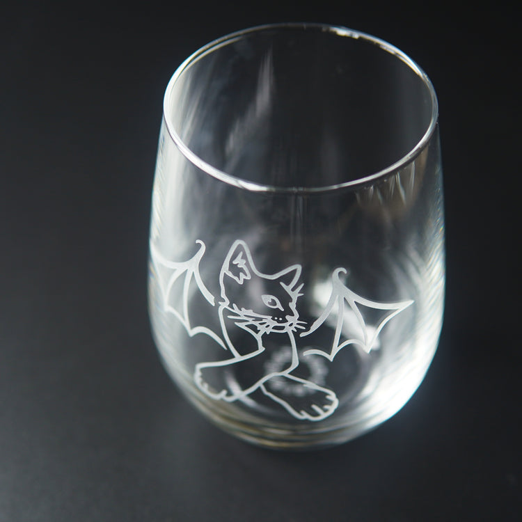 stemless wine glass engraved with a cat with crossed arms and bat wings. We're viewing the glass mostly from above