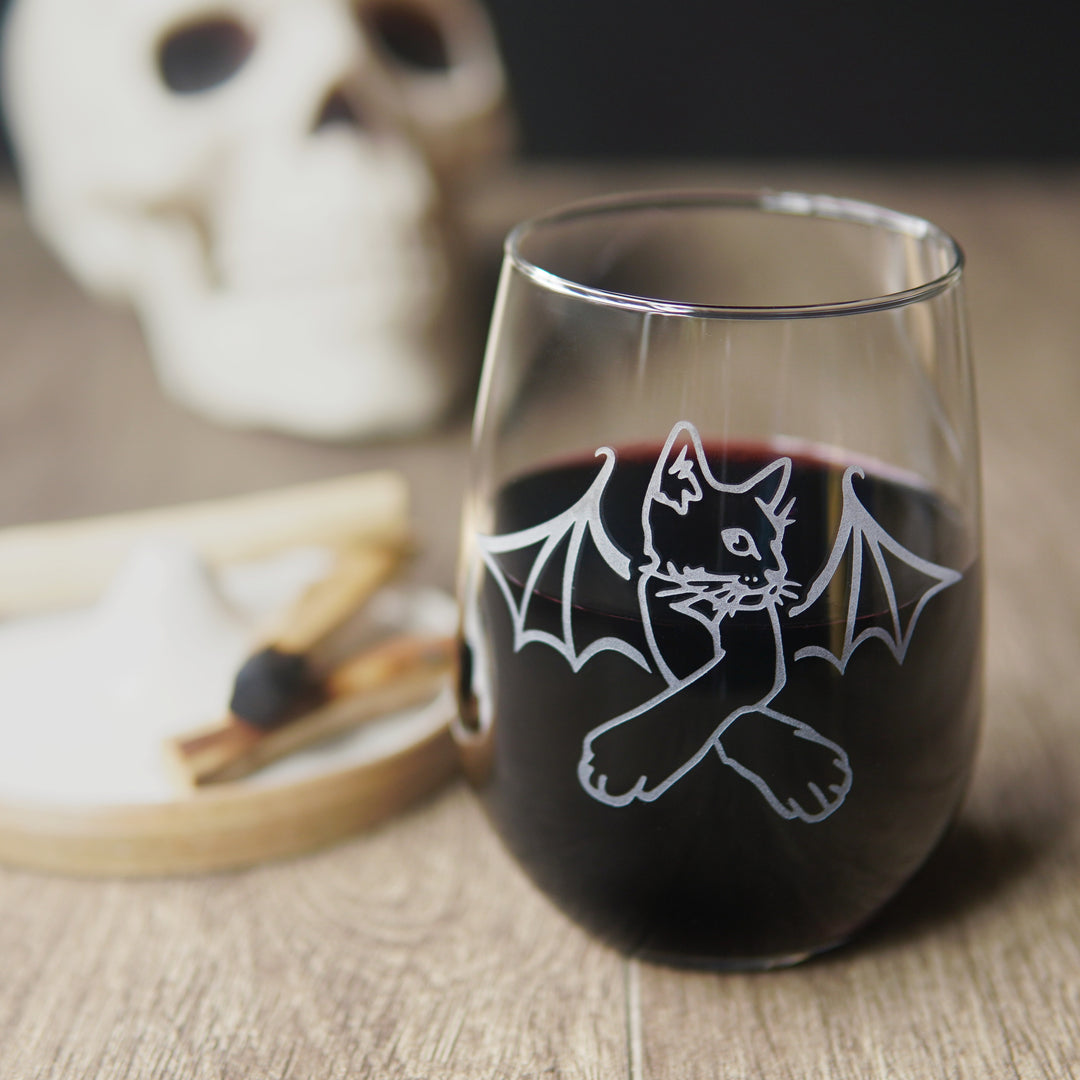 stemless wine glass engraved with a cat with crossed arms and bat wings. There is a human skull in the background, and a tray with palo santo sticks.