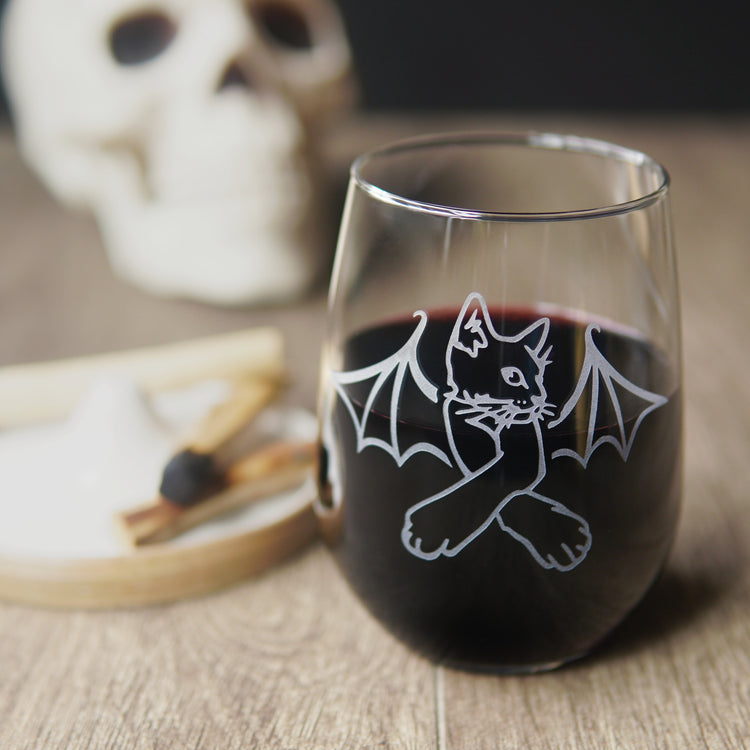 stemless wine glass engraved with a cat with crossed arms and bat wings. There is a human skull in the background, and a tray with palo santo sticks.