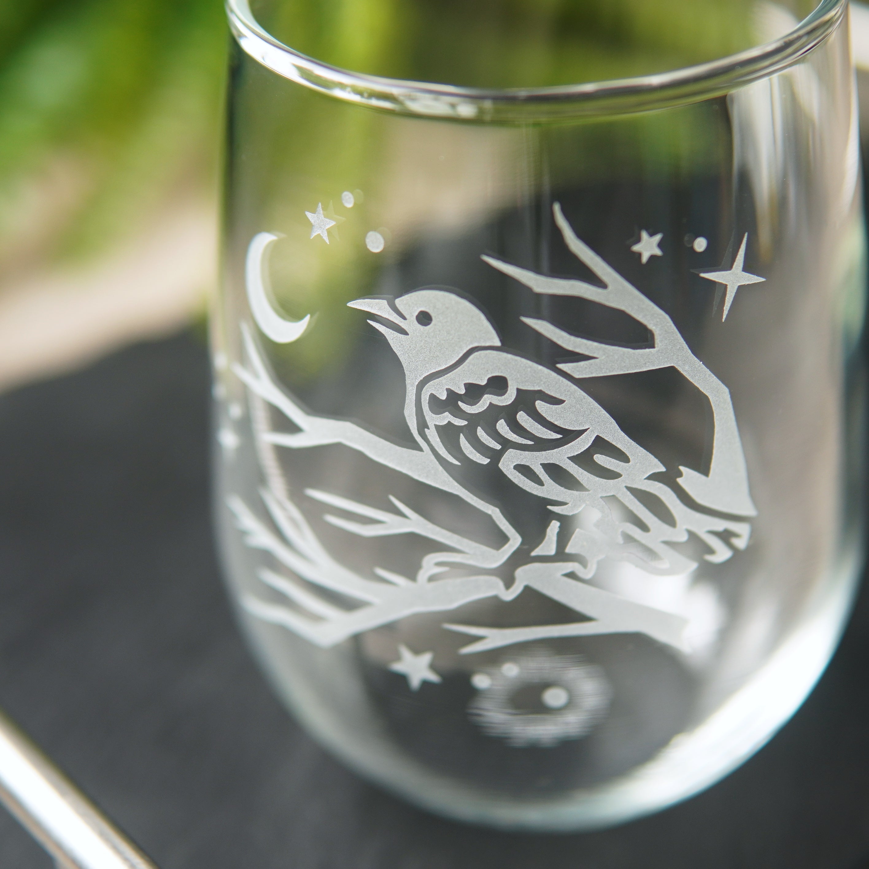 stemless wine glass etched with a crow in a tree, facing a crescent moon and stars, seen from close up.
