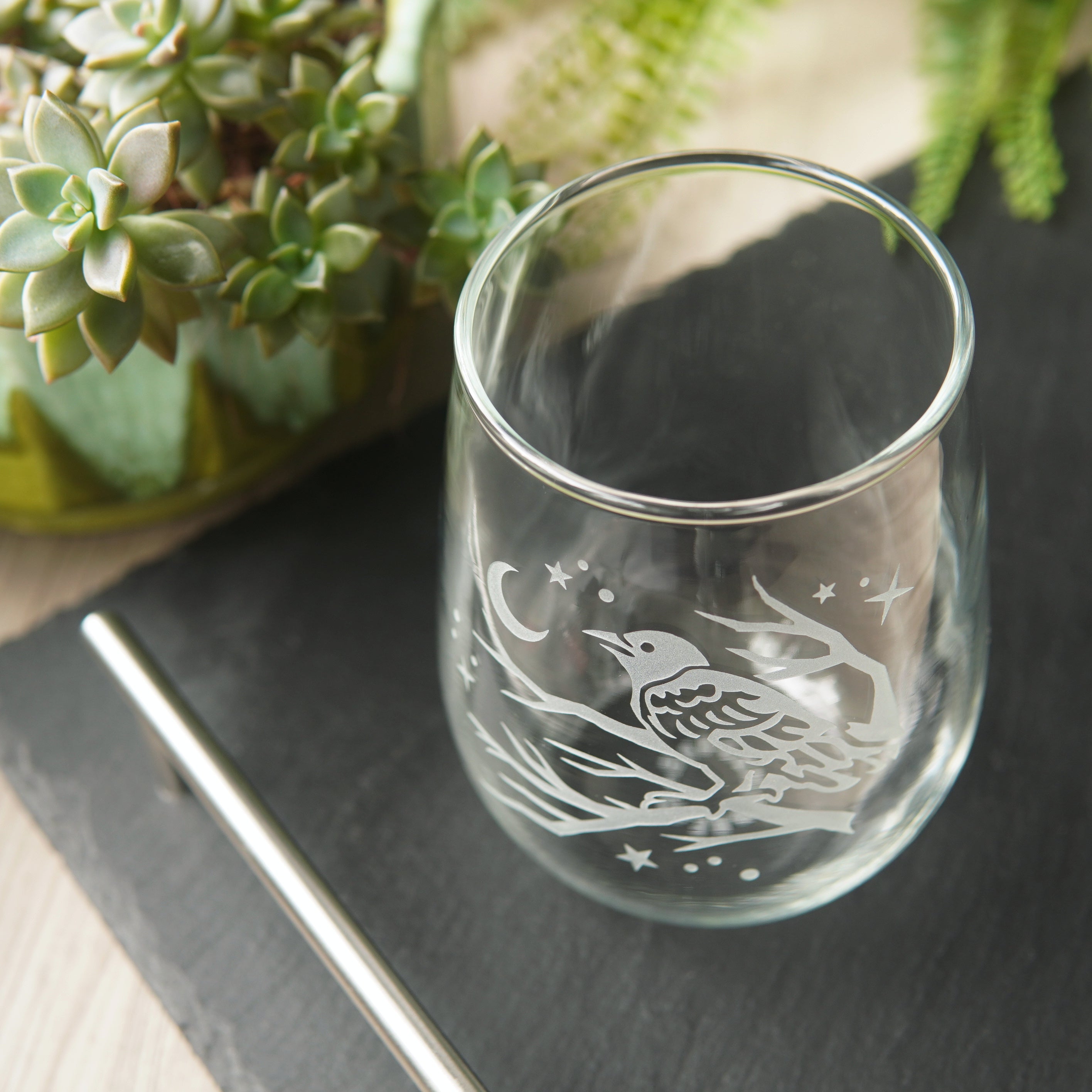 stemless wine glass etched with a crow in a tree, facing a crescent moon and stars. The glass is seen mostly from above, on a black slate tray, next to a succulent plant.
