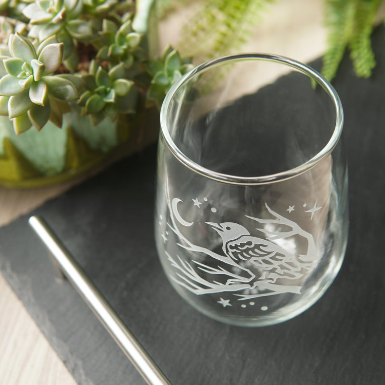 stemless wine glass etched with a crow in a tree, facing a crescent moon and stars. The glass is seen mostly from above, on a black slate tray, next to a succulent plant.
