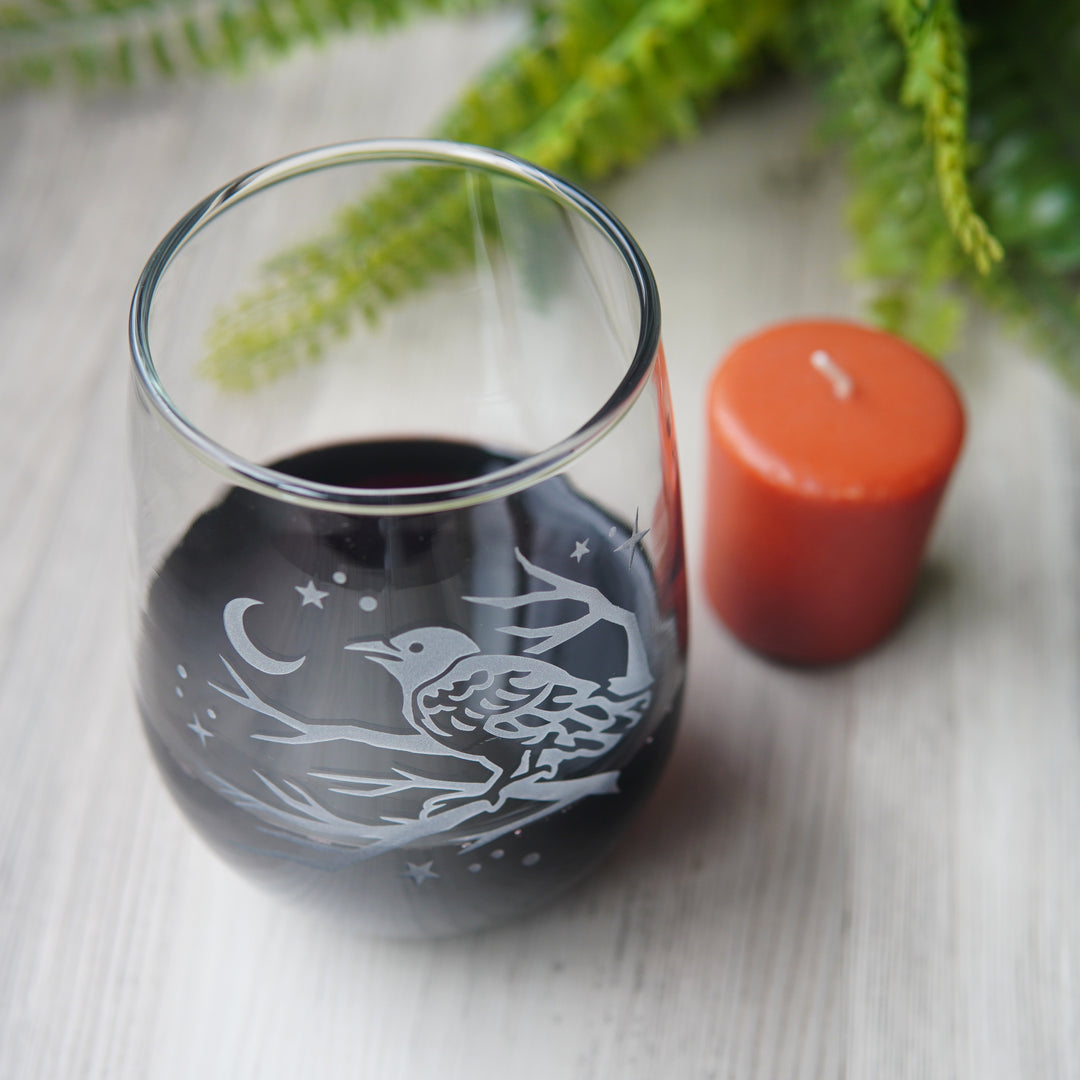 stemless wine glass etched with a crow in a tree, facing a crescent moon and stars, seen from above, next to a votive candle and fern.