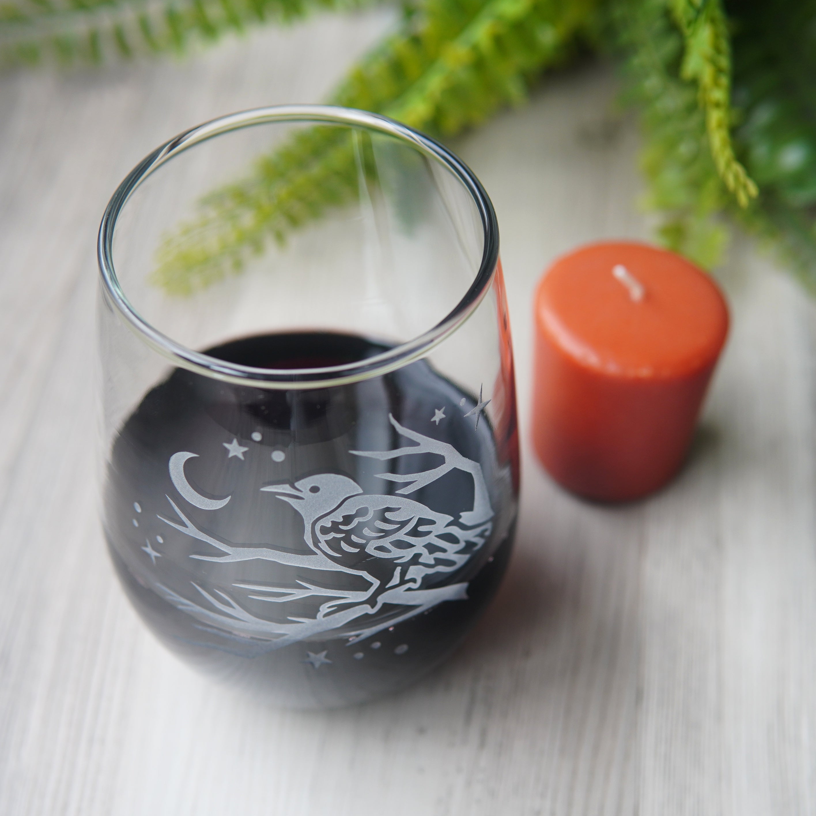 stemless wine glass etched with a crow in a tree, facing a crescent moon and stars, seen from above, next to a votive candle and fern.