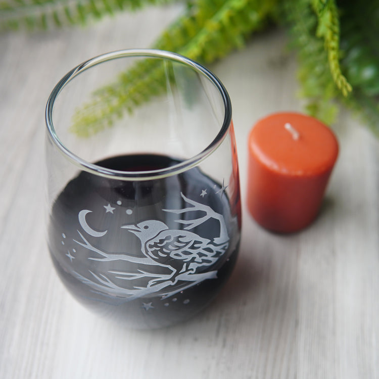 stemless wine glass etched with a crow in a tree, facing a crescent moon and stars, seen from above, next to a votive candle and fern.