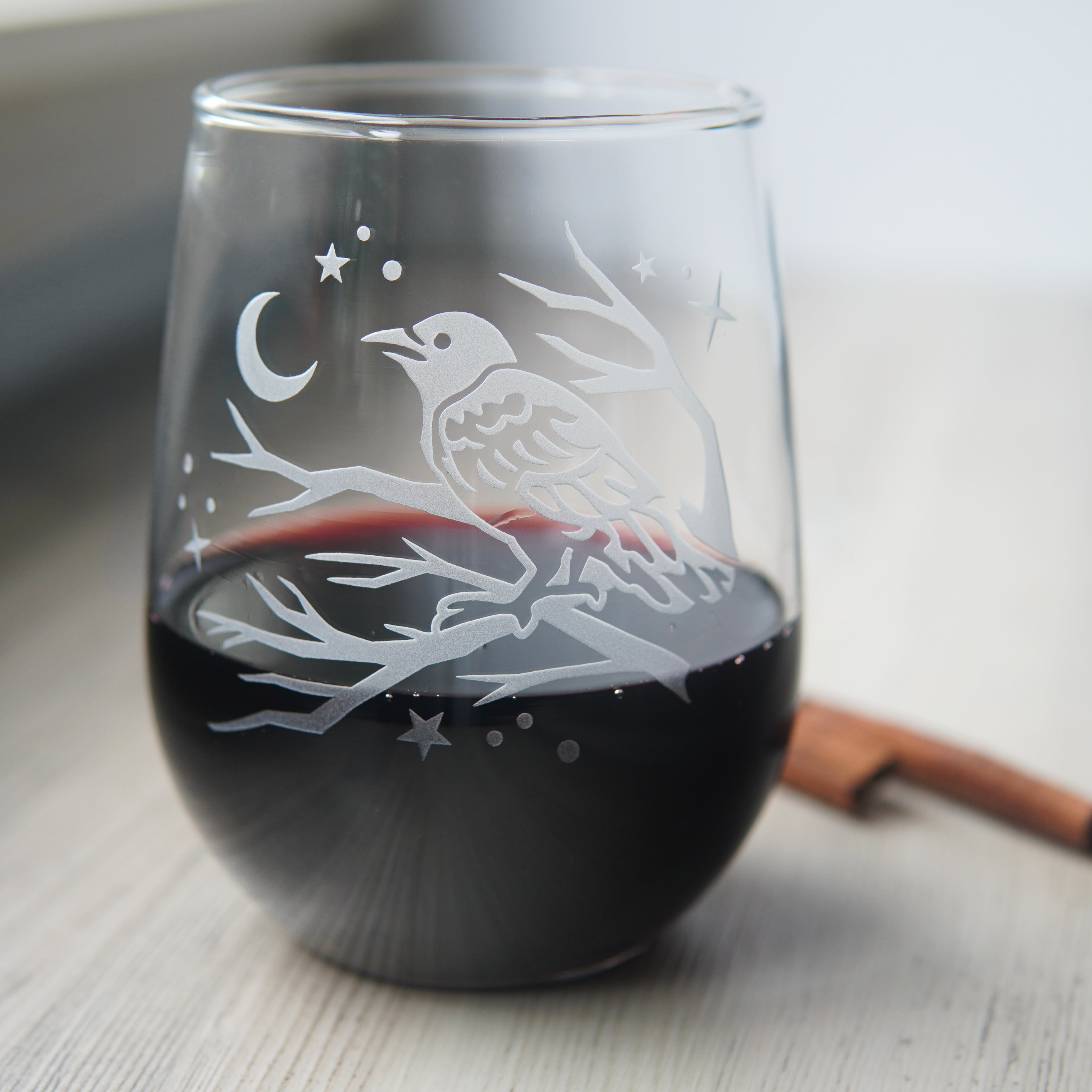 stemless wine glass etched with a crow in a tree, facing a crescent moon and stars. There is red wine in the glass.