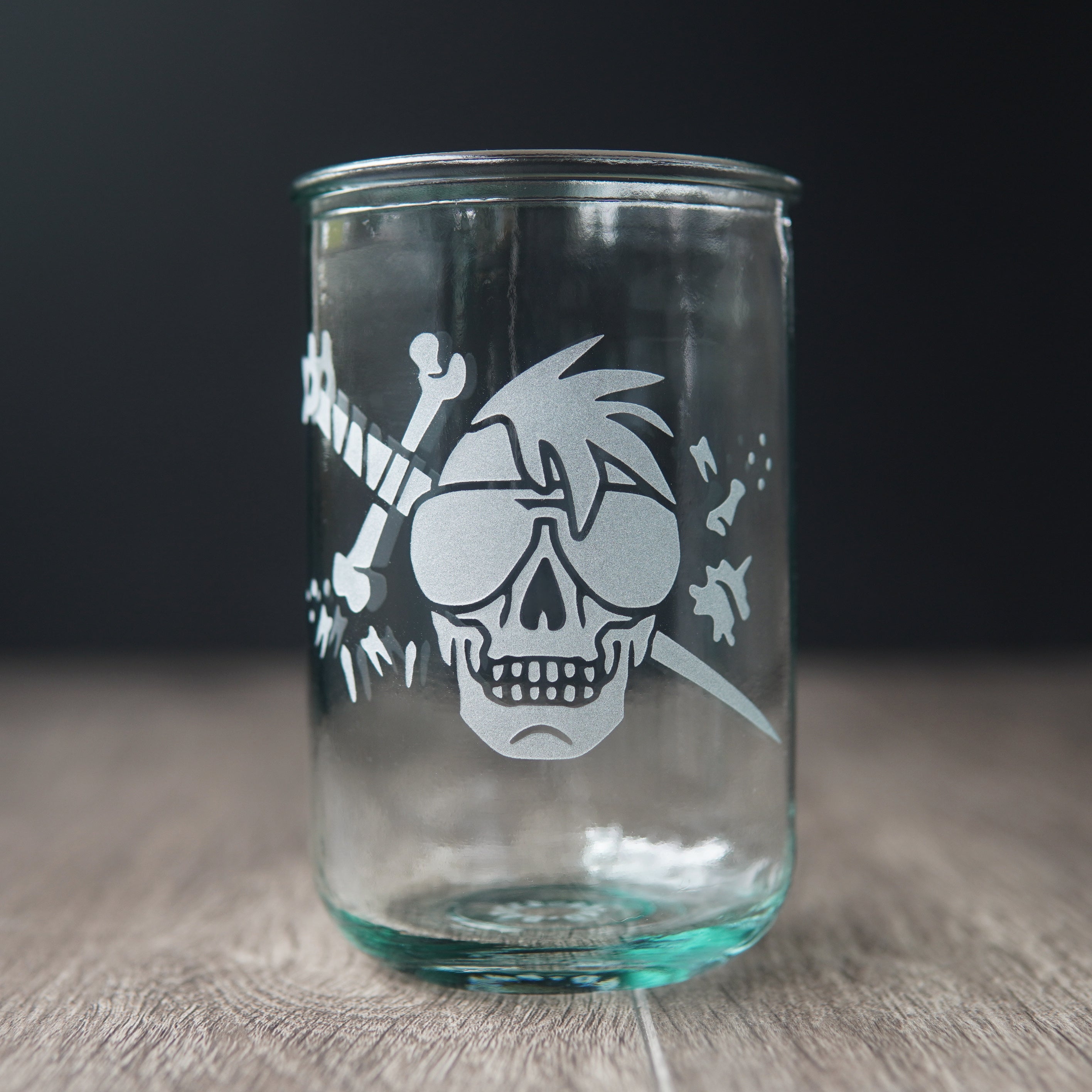 A straight-sided drinking glass with a slight green-blue tinge. The glass has a skull with a mohawk wearing sunglasses, with a sword and bones behind it.