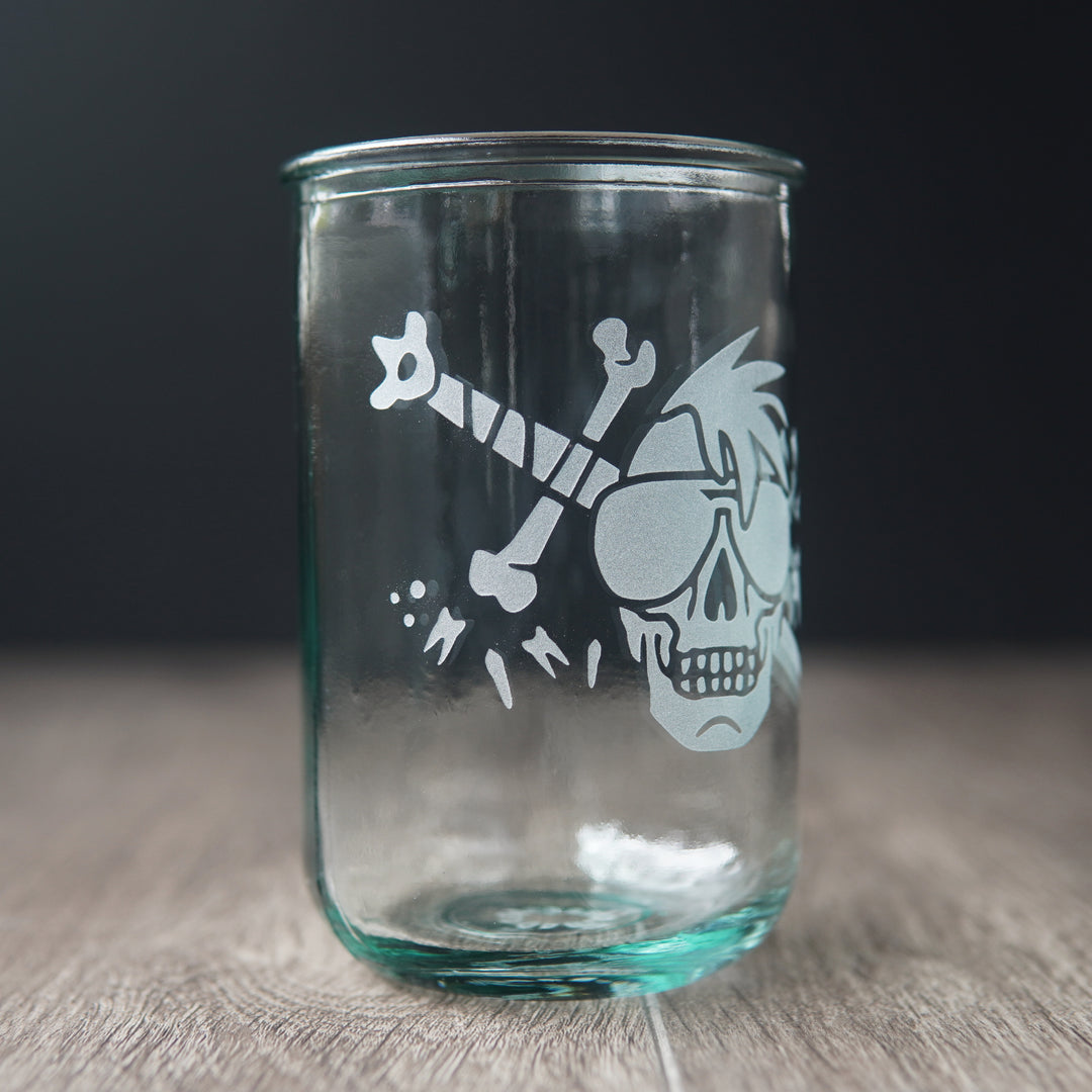 A straight-sided drinking glass with a slight green-blue tinge. The glass has a skull with a mohawk wearing sunglasses, with a sword and teeth behind it.