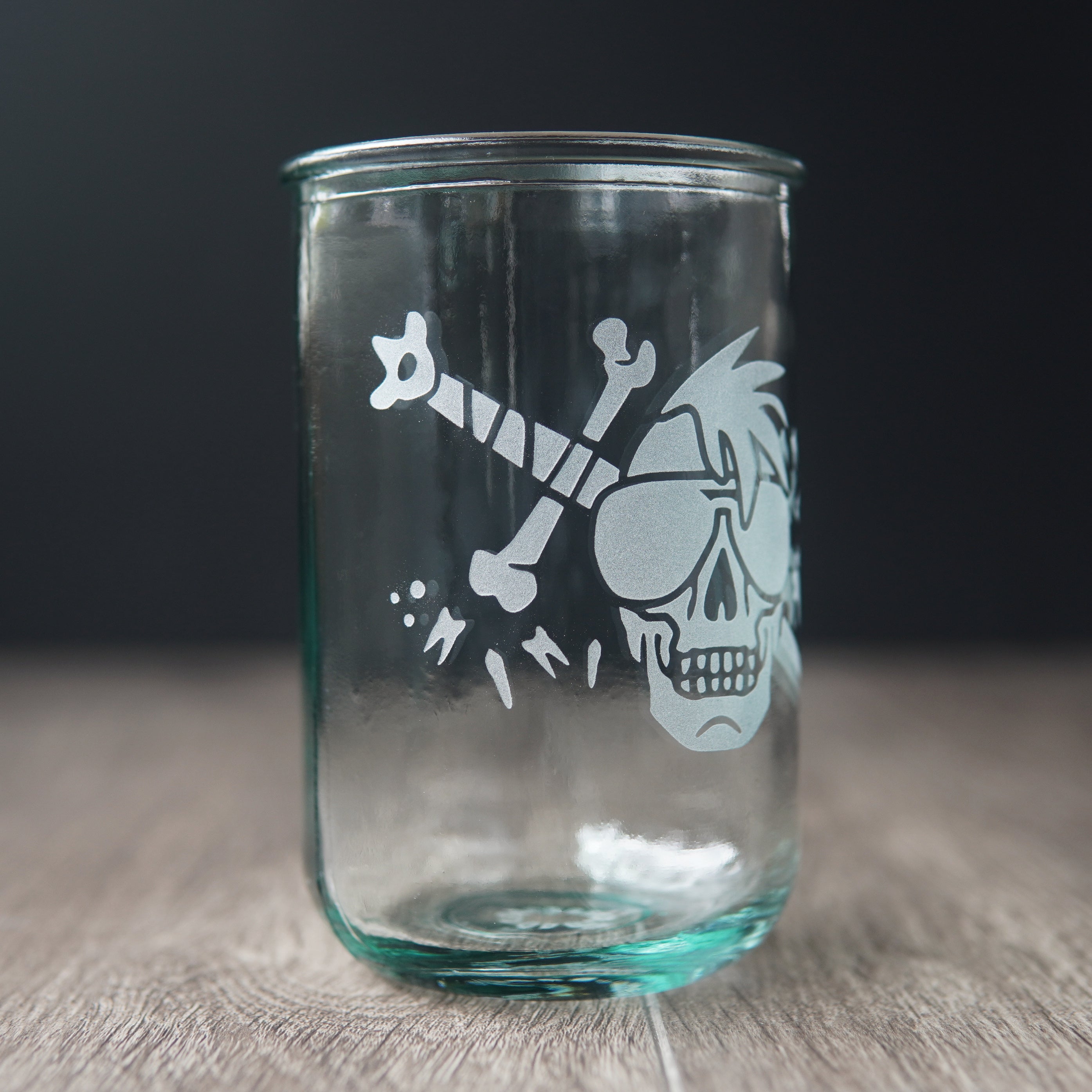 A straight-sided drinking glass with a slight green-blue tinge. The glass has a skull with a mohawk wearing sunglasses, with a sword and teeth behind it.