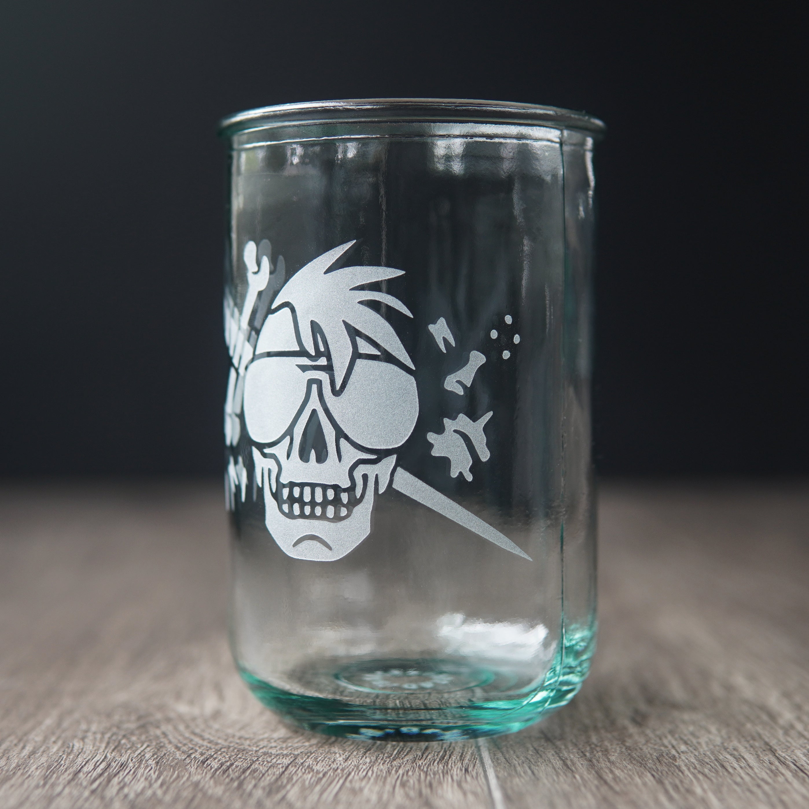 A straight-sided drinking glass with a slight green-blue tinge. The glass has a skull with a mohawk wearing sunglasses, with a sword and bones behind it.