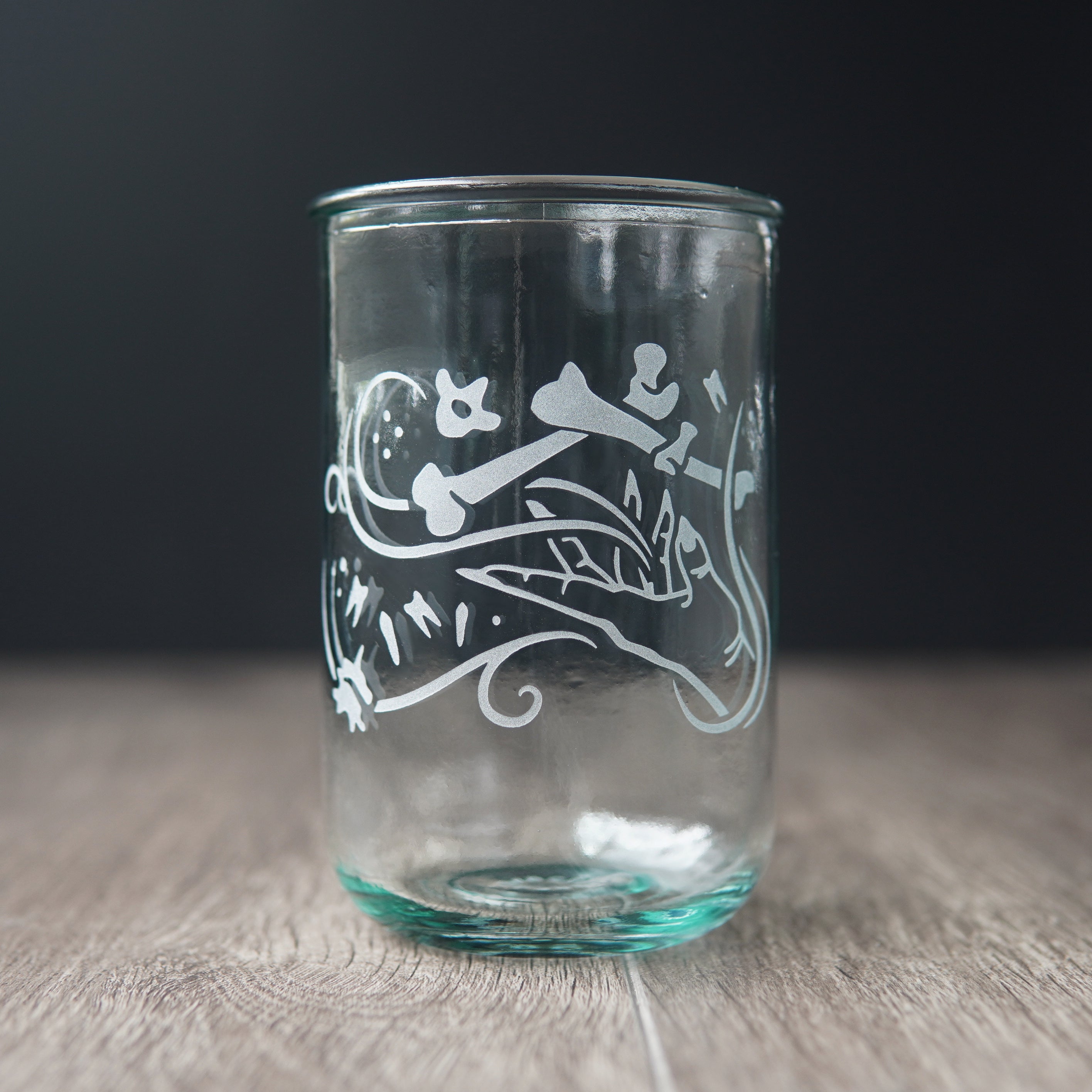A straight-sided drinking glass with a slight green-blue tinge. The glass has  a hand and assorted bones on it, with swirly spirals suggesting magic and movement between the hand and bones.
