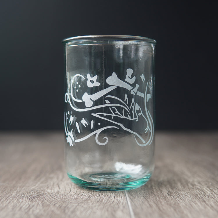 A straight-sided drinking glass with a slight green-blue tinge. The glass has  a hand and assorted bones on it, with swirly spirals suggesting magic and movement between the hand and bones.