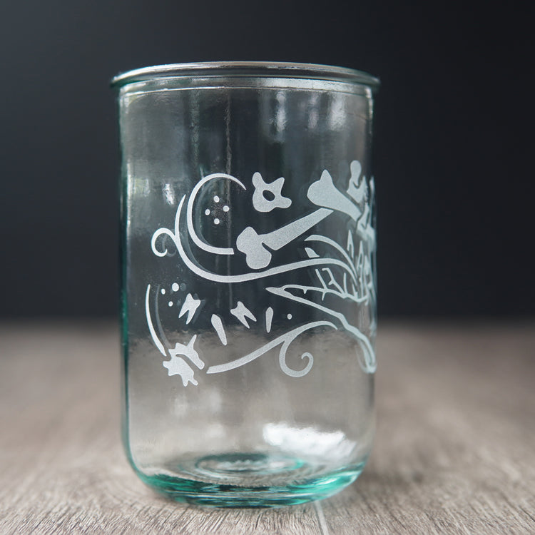 A straight-sided drinking glass with a slight green-blue tinge. The glass has  a hand and assorted bones on it, with swirly spirals suggesting magic and movement between the hand and bones.