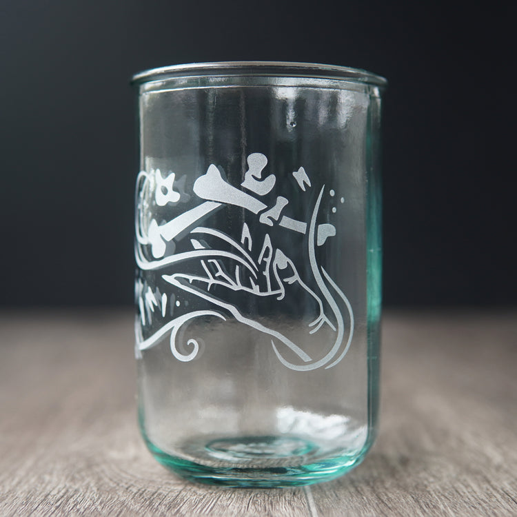 A straight-sided drinking glass with a slight green-blue tinge. The glass has  a hand and assorted bones on it, with swirly spirals suggesting magic and movement between the hand and bones.