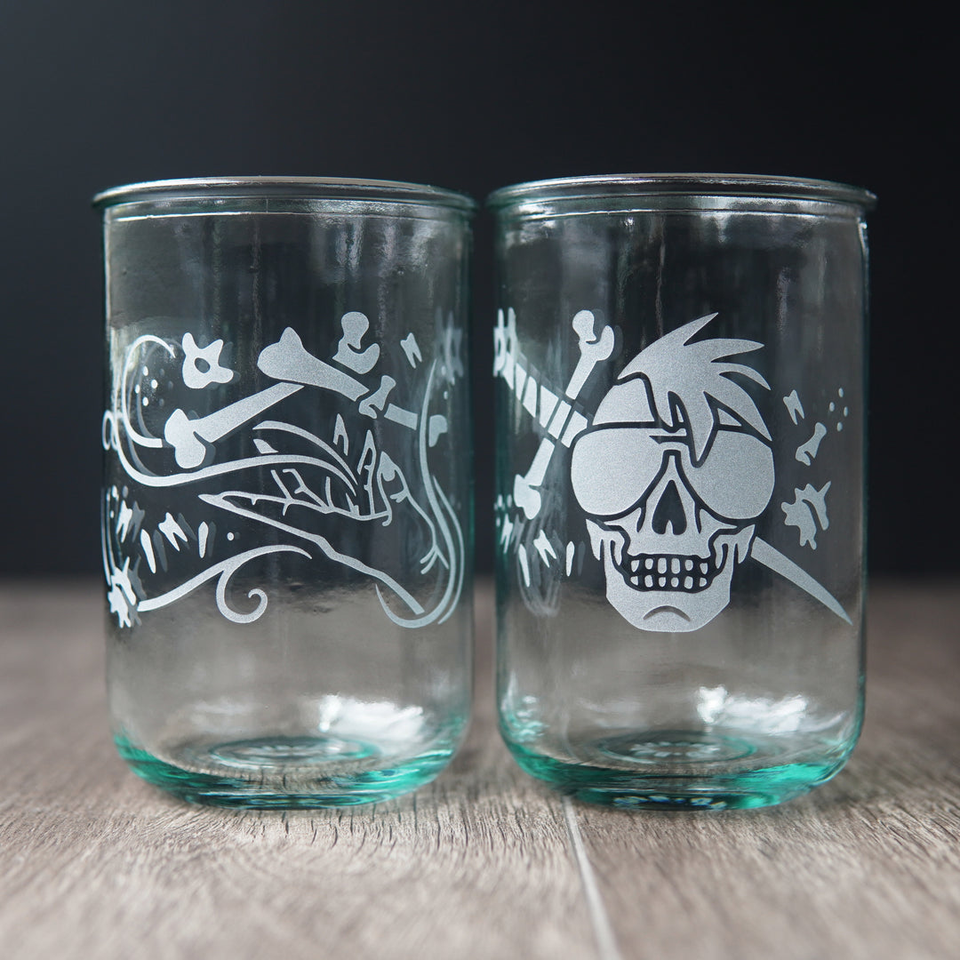 Two straight-sided drinking glasses with a slight green-blue tinge. One has a hand and assorted bones on it. The second glass has a skull with a mohawk wearing sunglasses with a sword and bones behind it.
