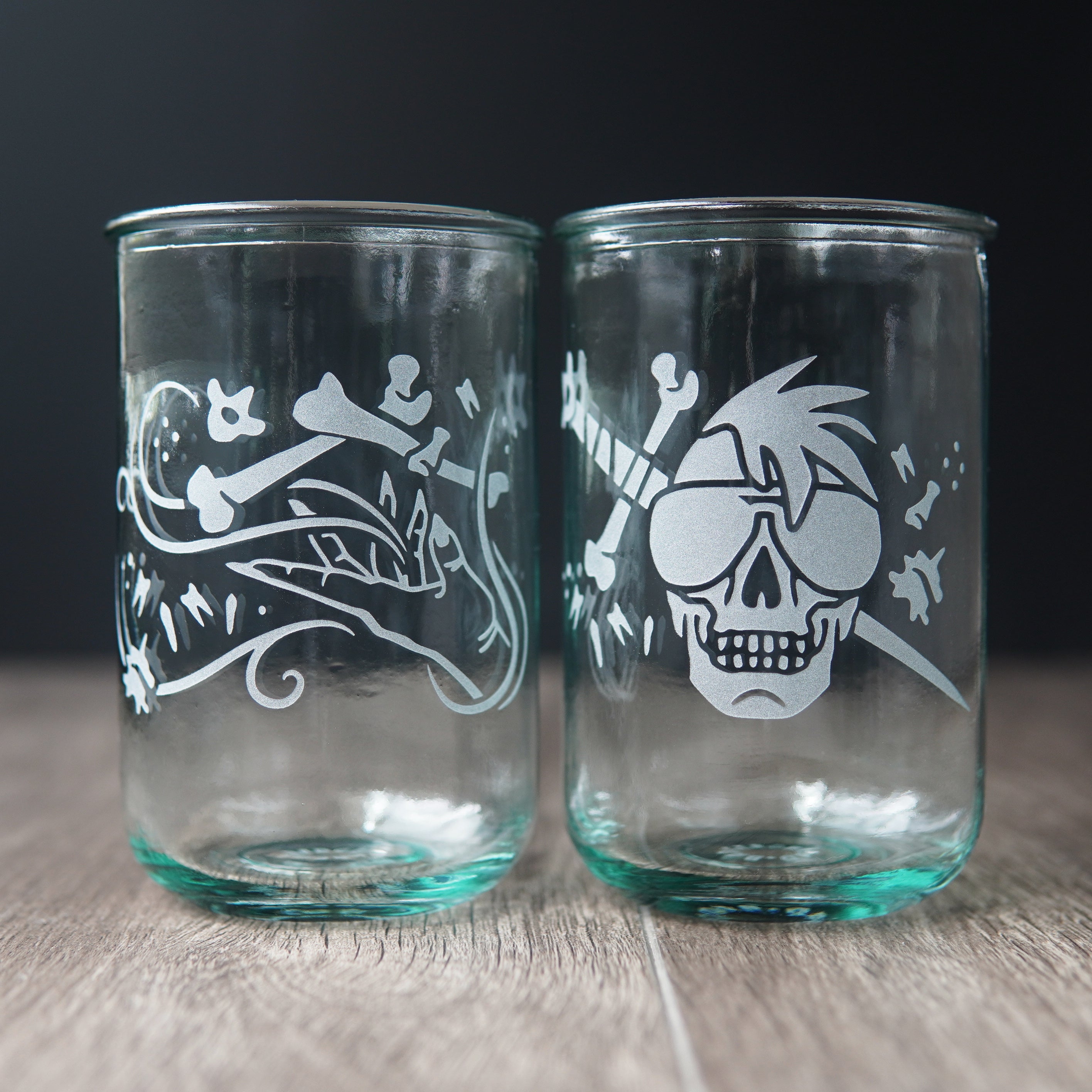 Two straight-sided drinking glasses with a slight green-blue tinge. One has a hand and assorted bones on it. The second glass has a skull with a mohawk wearing sunglasses with a sword and bones behind it.