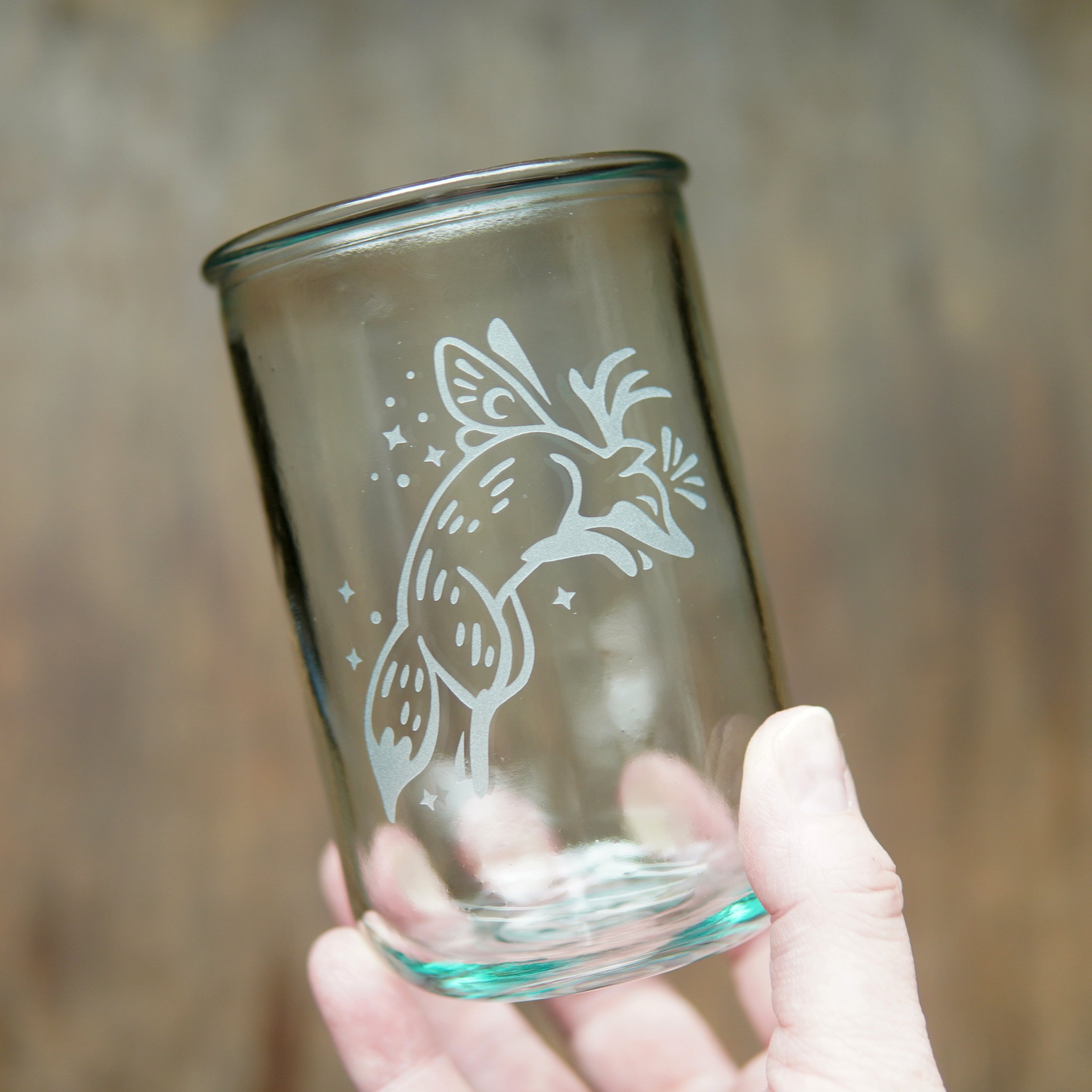 Fox with fairy wings and antlers etched onto a tall glass, held by a hand