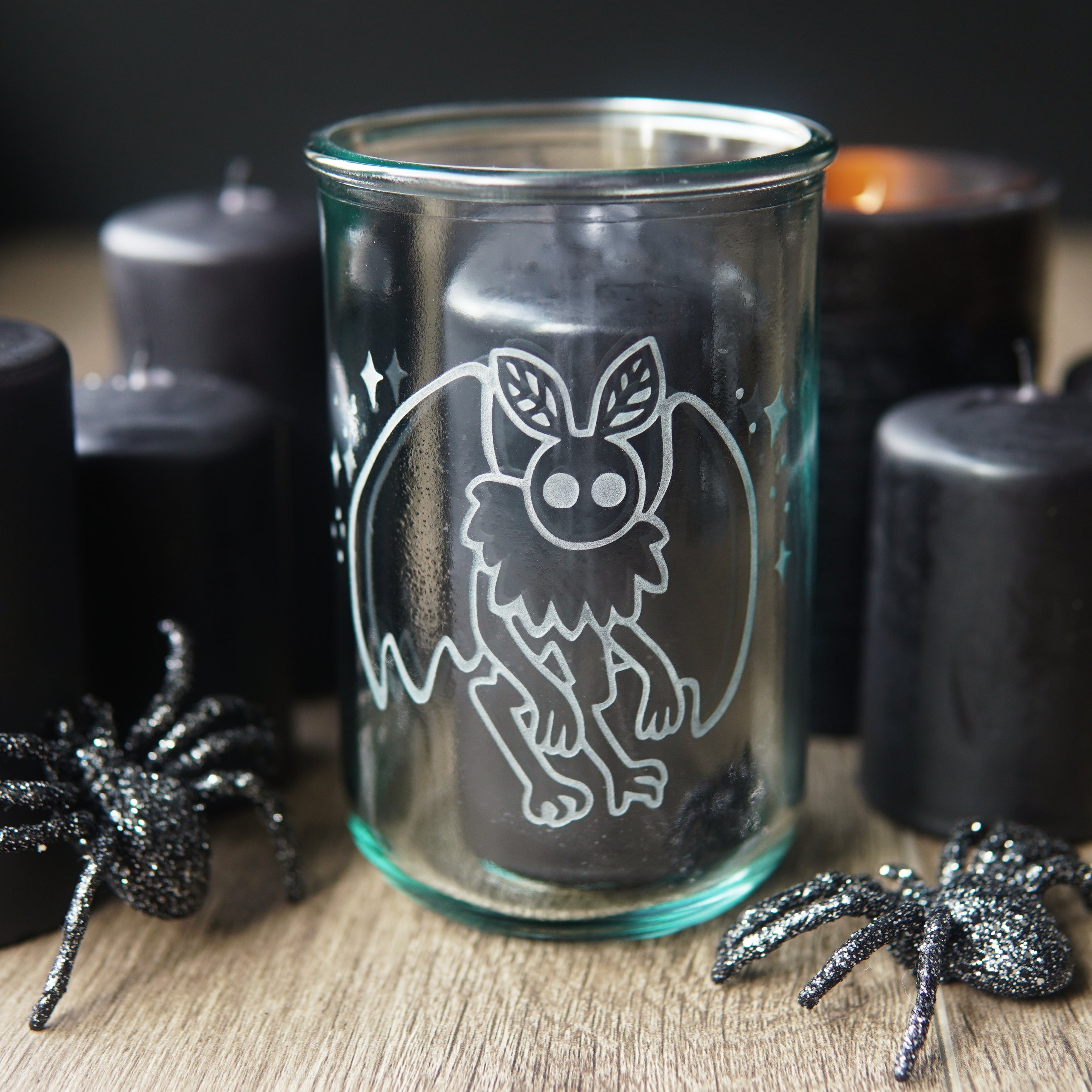 mothman etched onto a tall glass, with black pillar candles and plastic spiders