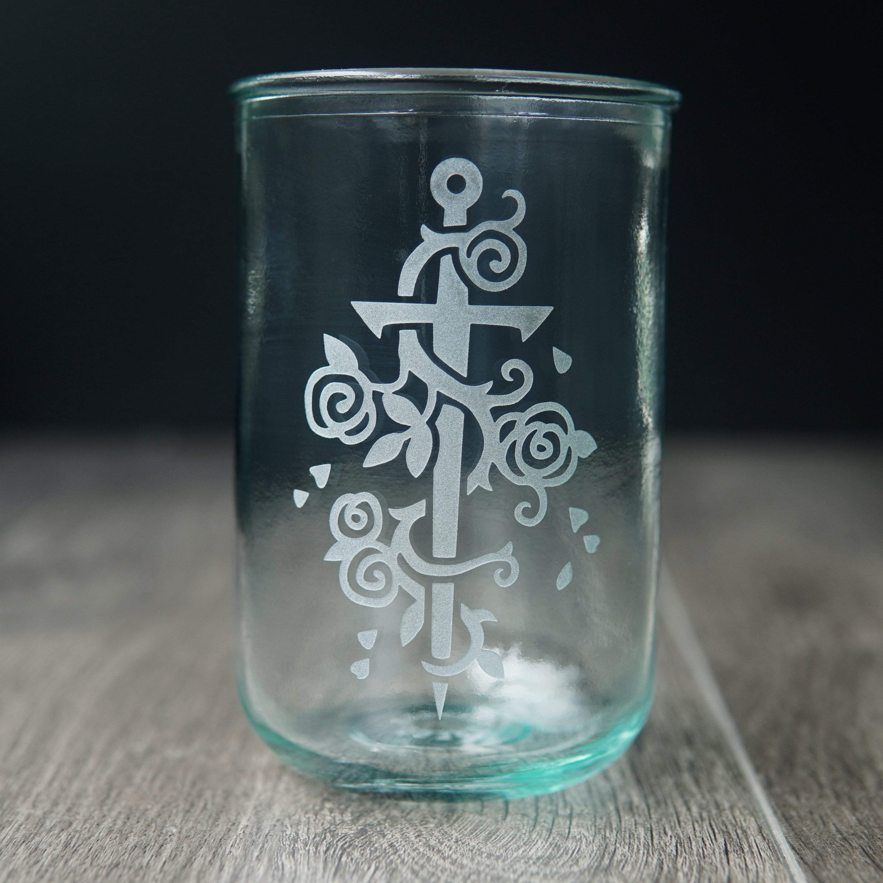 Sword wrapped with a rose vine engraved onto a tall drinking glass. The glass has a slight bluegreen tinge.