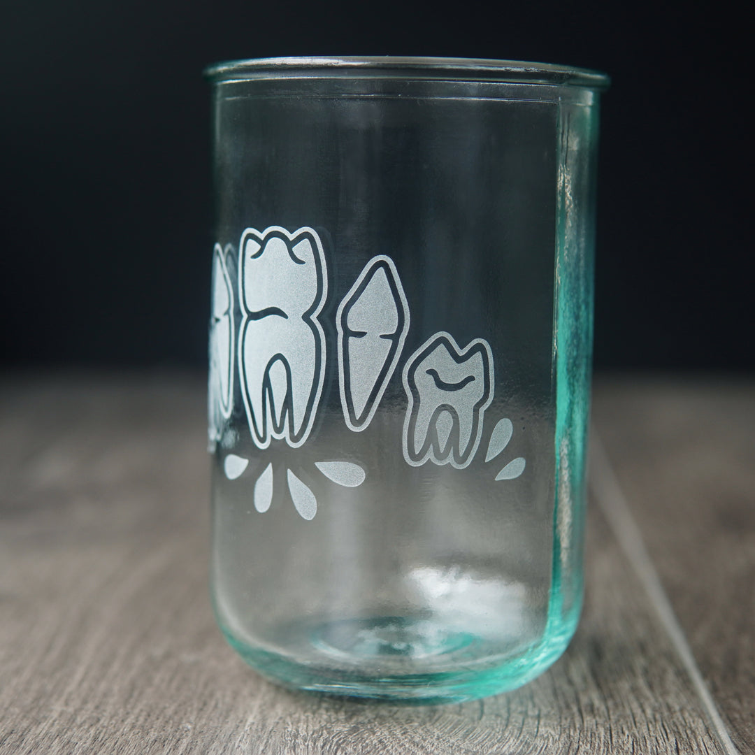 Four teeth with blood droplets engraved onto a tall drinking glass. The glass has a slight bluegreen tinge and is viewed slightly from the side.
