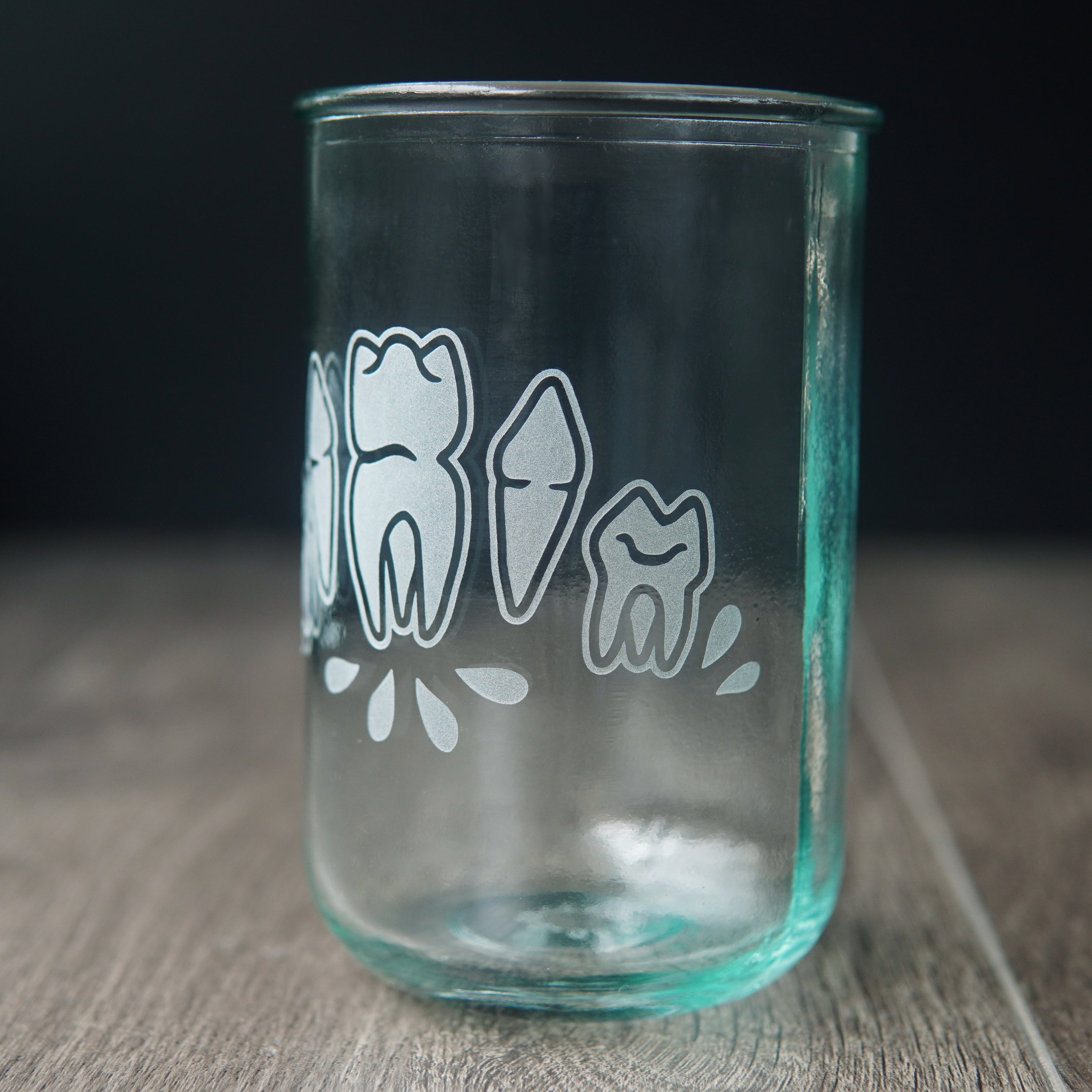 Four teeth with blood droplets engraved onto a tall drinking glass. The glass has a slight bluegreen tinge and is viewed slightly from the side.