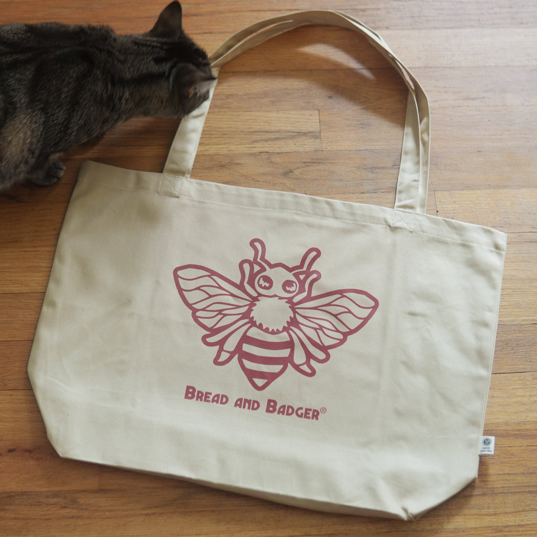 Beige tote bag with red bee design and 'Bread and Badger' text on a wood floor, with a cat sniffing the bag.