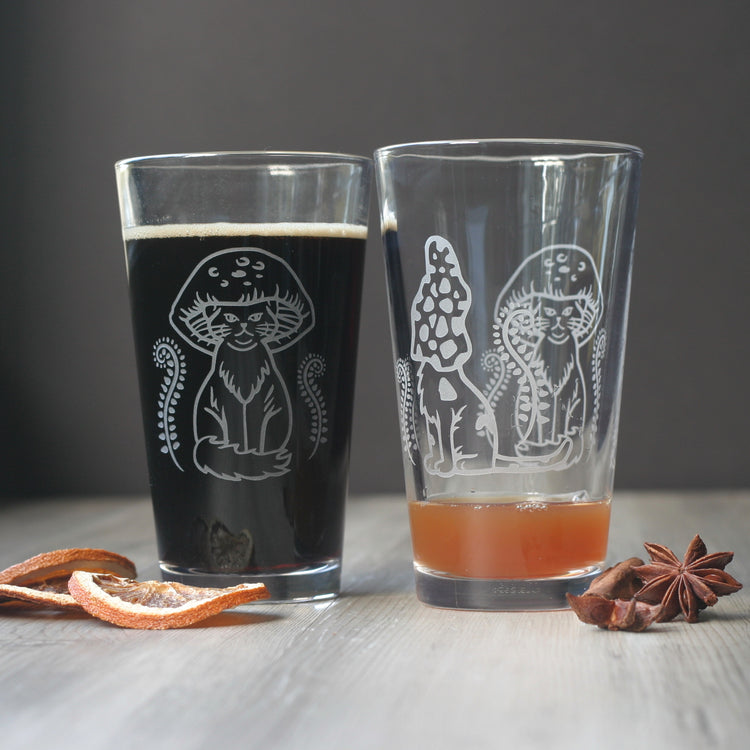 pair of pint glasses, engraved with two different cat designs wearing mushroom caps on their heads