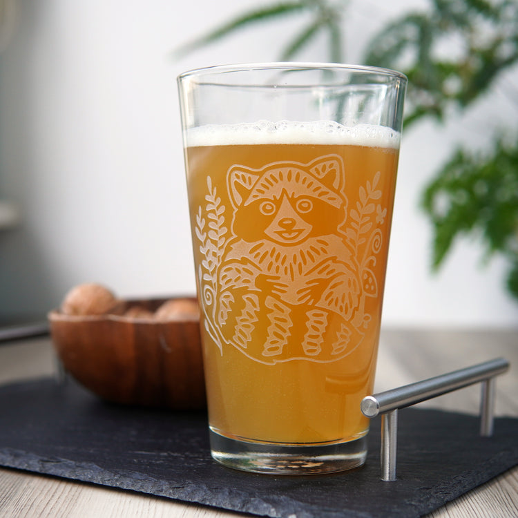 etched pint glass engraved with a cute, smiling raccoon, full of hazy beer